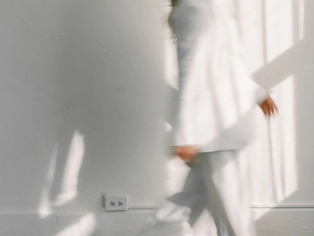Blurry image of a person dressed in white standing near a wall with electrical outlets, with sunlight casting shadows.