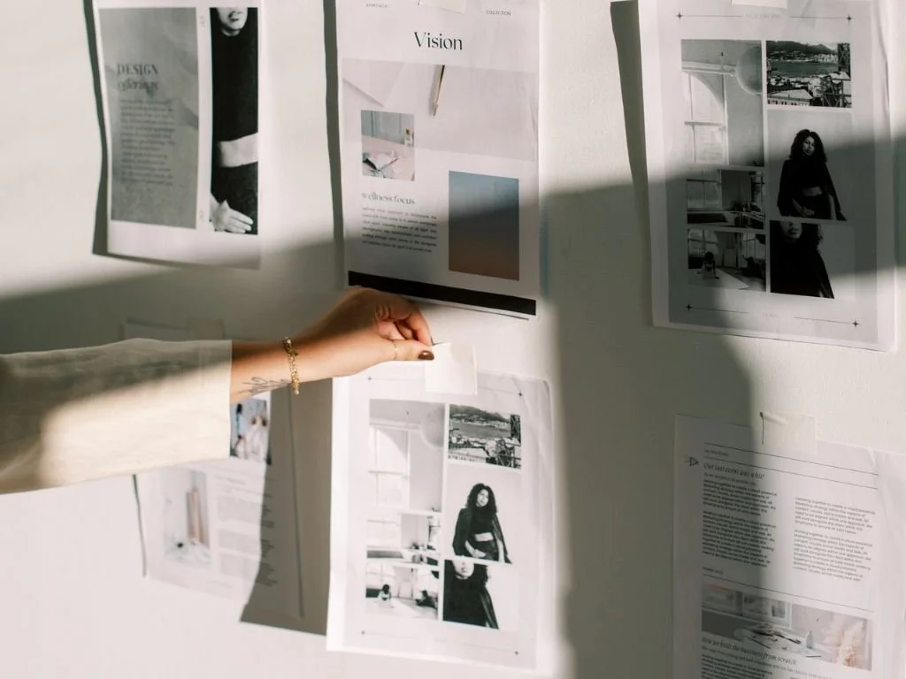 Person pinning a photo on a wall filled with black-and-white magazine pages and photos, with sunlight casting shadows.