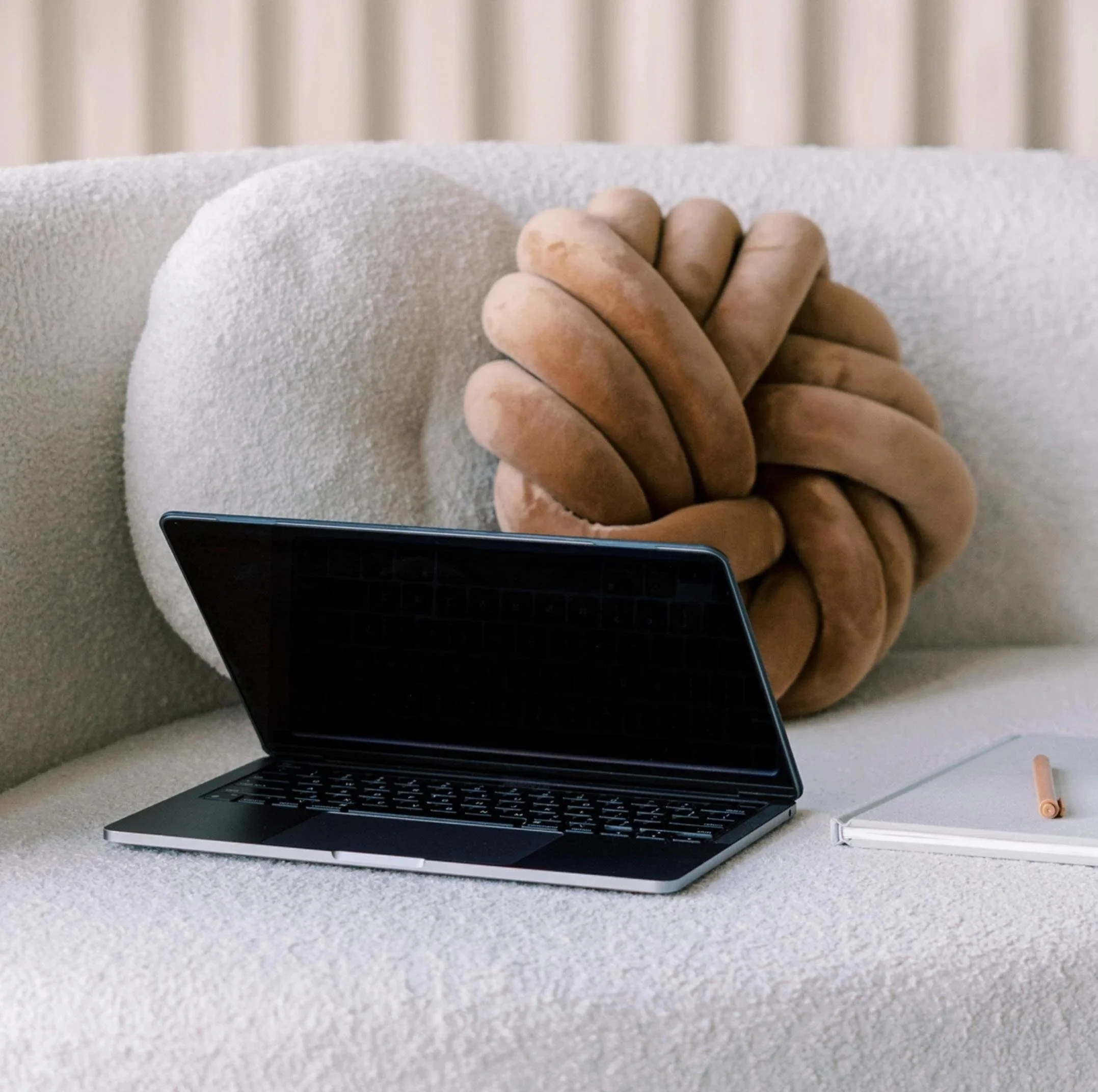 A black laptop, a white notebook with a gold pen, and a brown knot-shaped pillow on a beige sofa with light-colored curtains in the background.