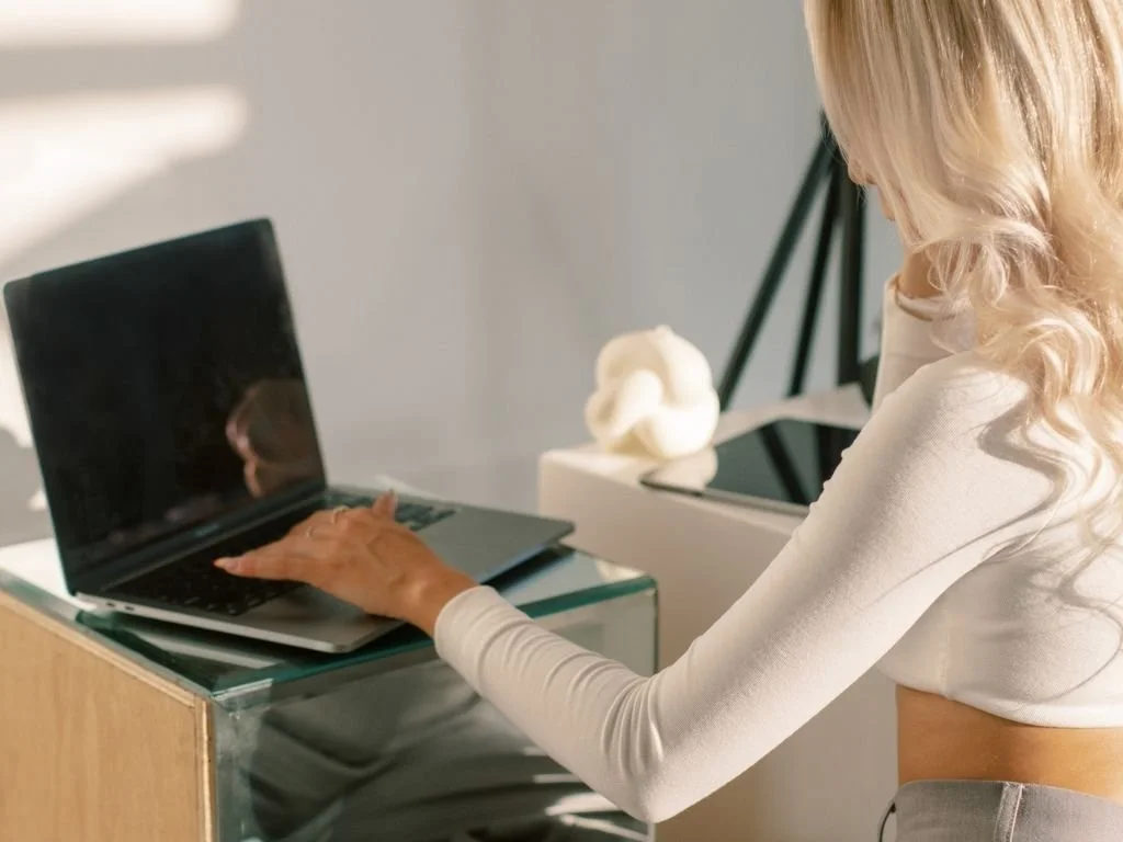 A woman with long blonde curly hair, wearing a white long-sleeve top, sitting at a desk and using a laptop.