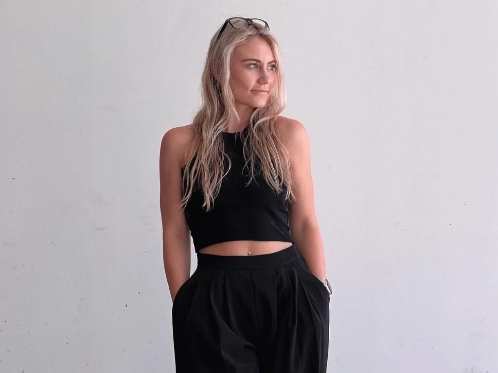 A young woman with long blonde hair, wearing glasses on her head, a black sleeveless crop top, and black high-waisted pants, stands against a plain white wall, looking to her left with her hands in her pockets.