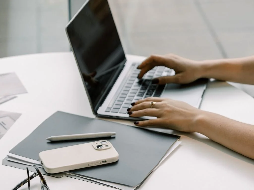 Person typing on a laptop with a smartphone, notebook, pen, and eyeglasses on a white desk.
