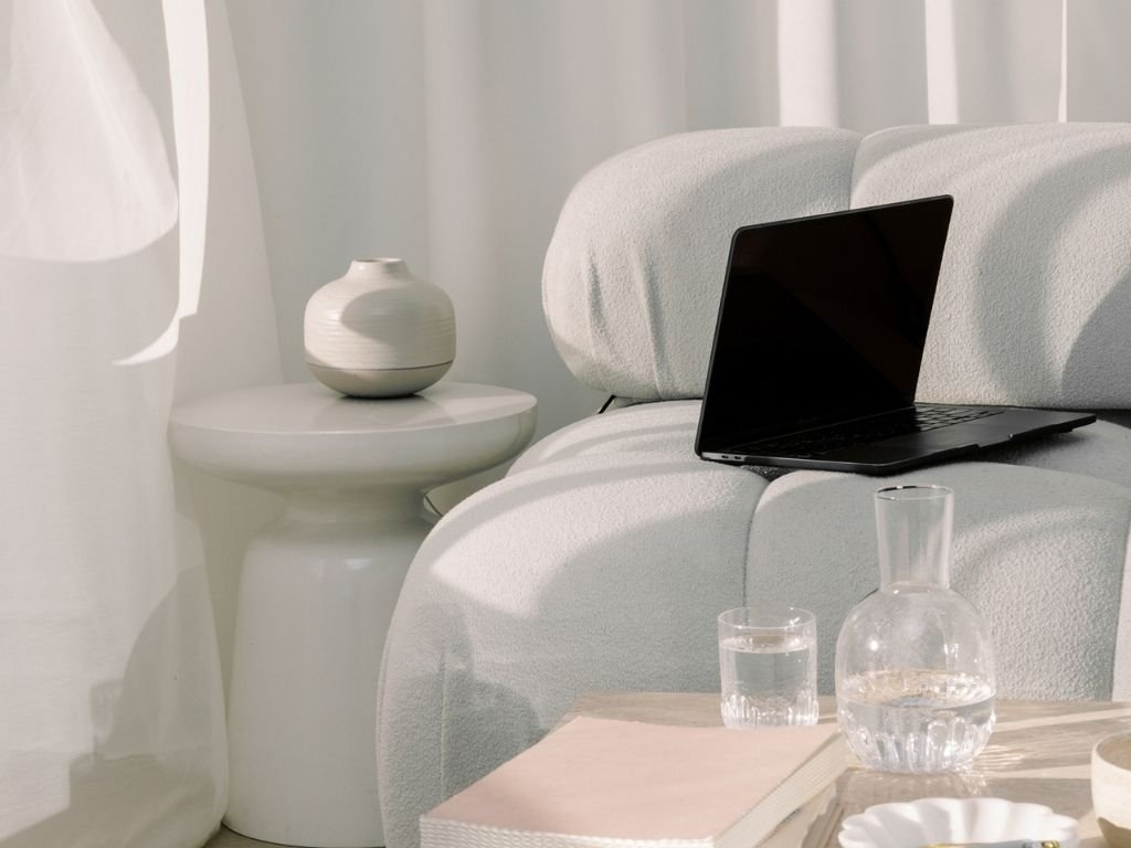 A modern living room corner with a white couch, a round side table with a white vase, a laptop on the couch, a glass pitcher, a glass of water, a book, and decorative items.