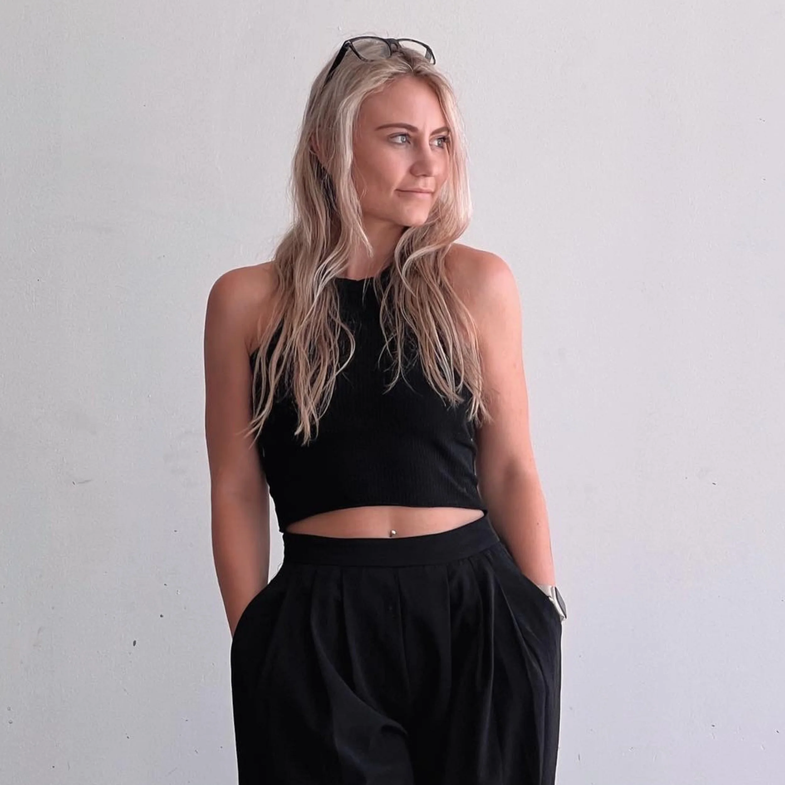 A young woman with long blond wavy hair, wearing glasses on her head, a black sleeveless crop top, and black pants, stands against a plain light gray wall, looking to her left.