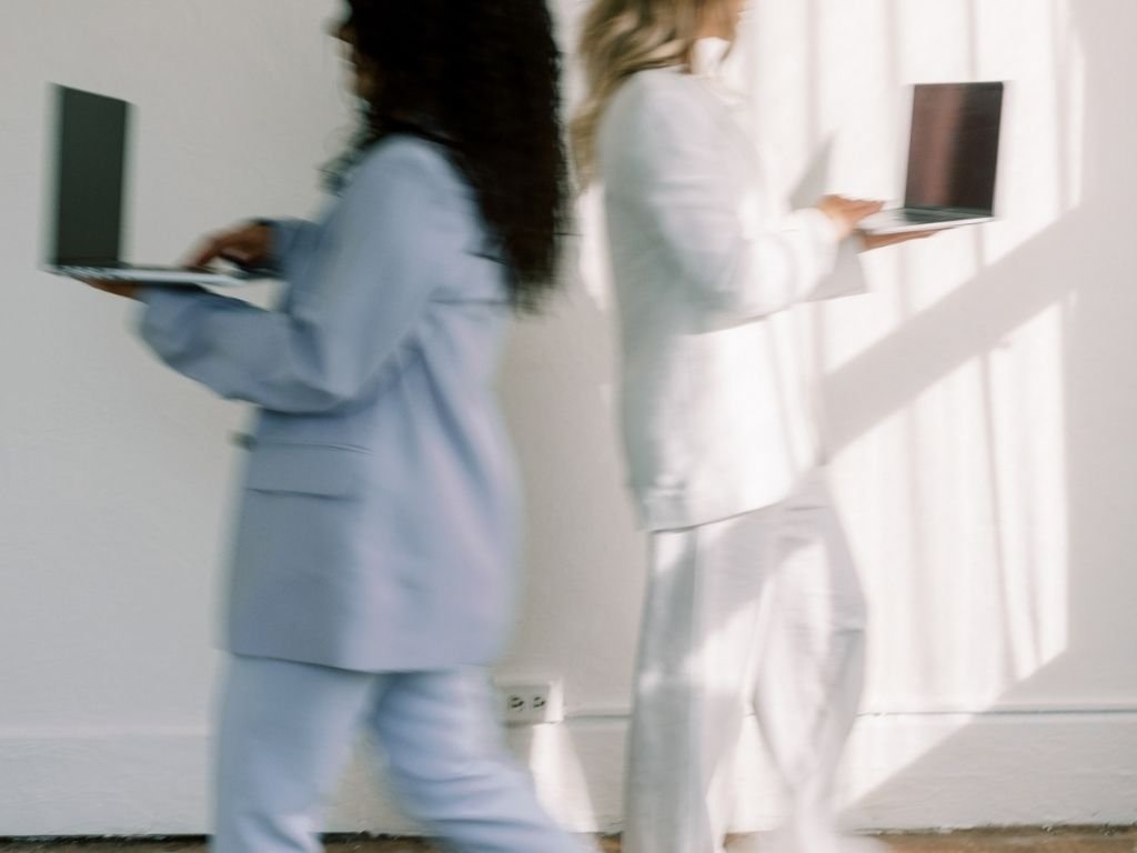 Two women in white suits walking side by side while using laptops in a bright room.