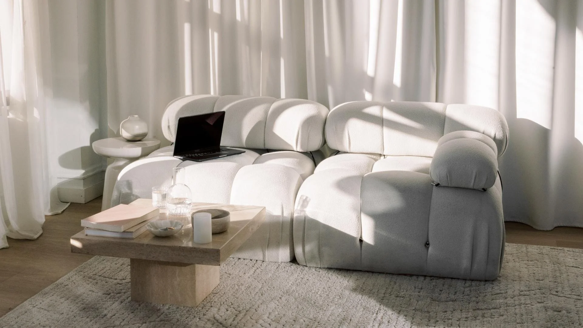 Living room with a white sofa, a small side table with a vase, a laptop, and a coffee table with books, a candle, and decorative items, with sunlight and curtains in the background.