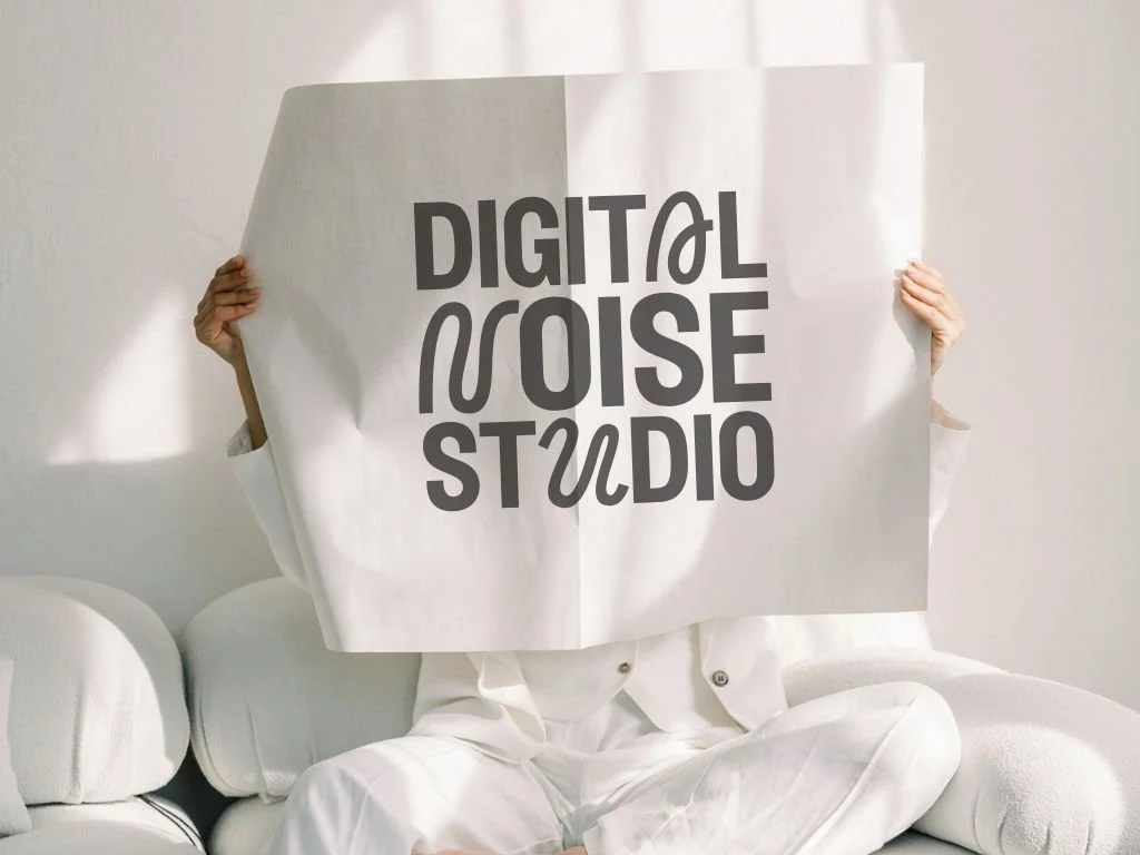 Person in white pajamas sitting on a white sofa holding a large sign that reads 'Digital Noise Studio' in black letters.