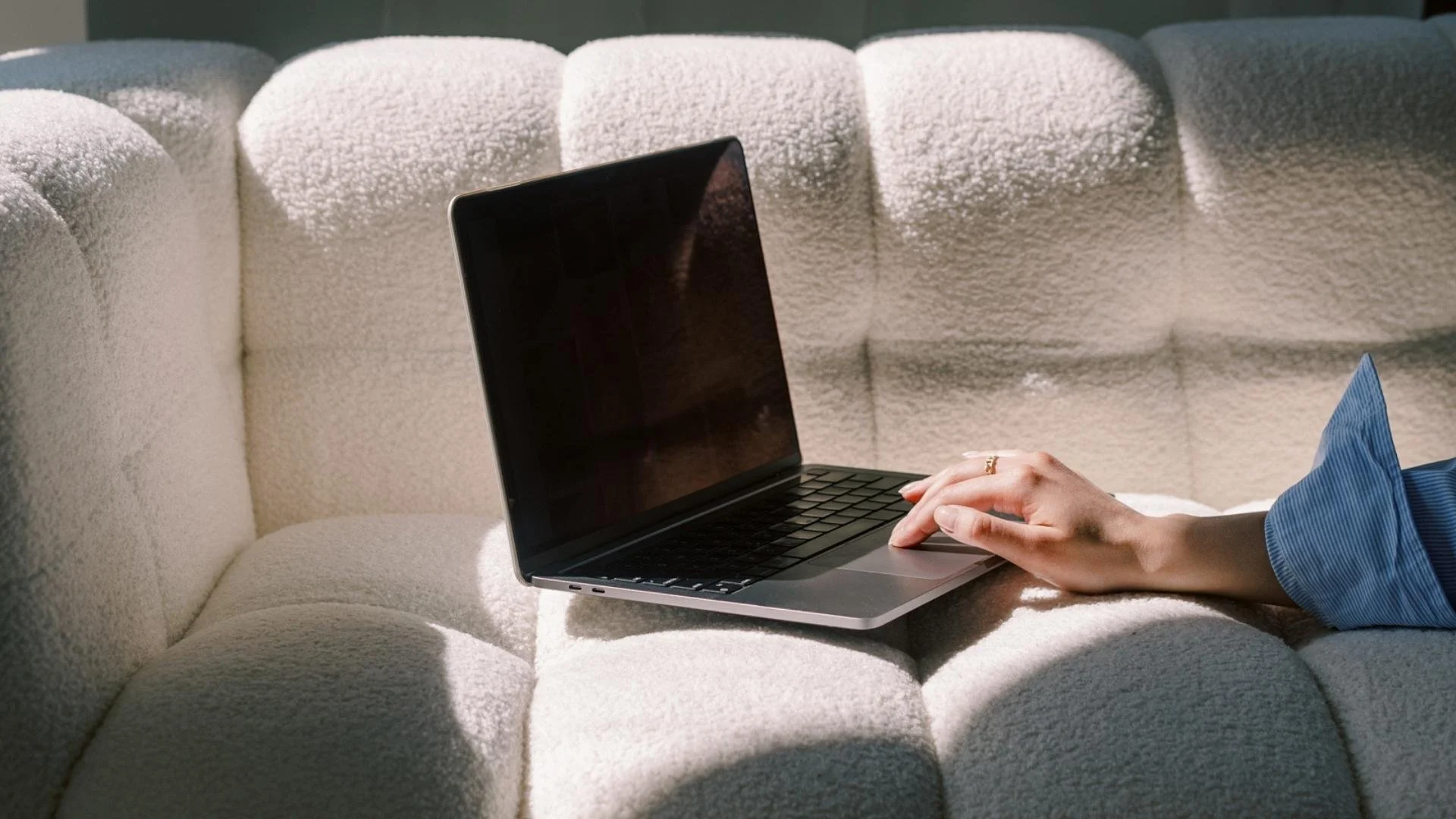 A person's hand with a gold ring on the finger, resting on a laptop on a textured cream-colored sofa.