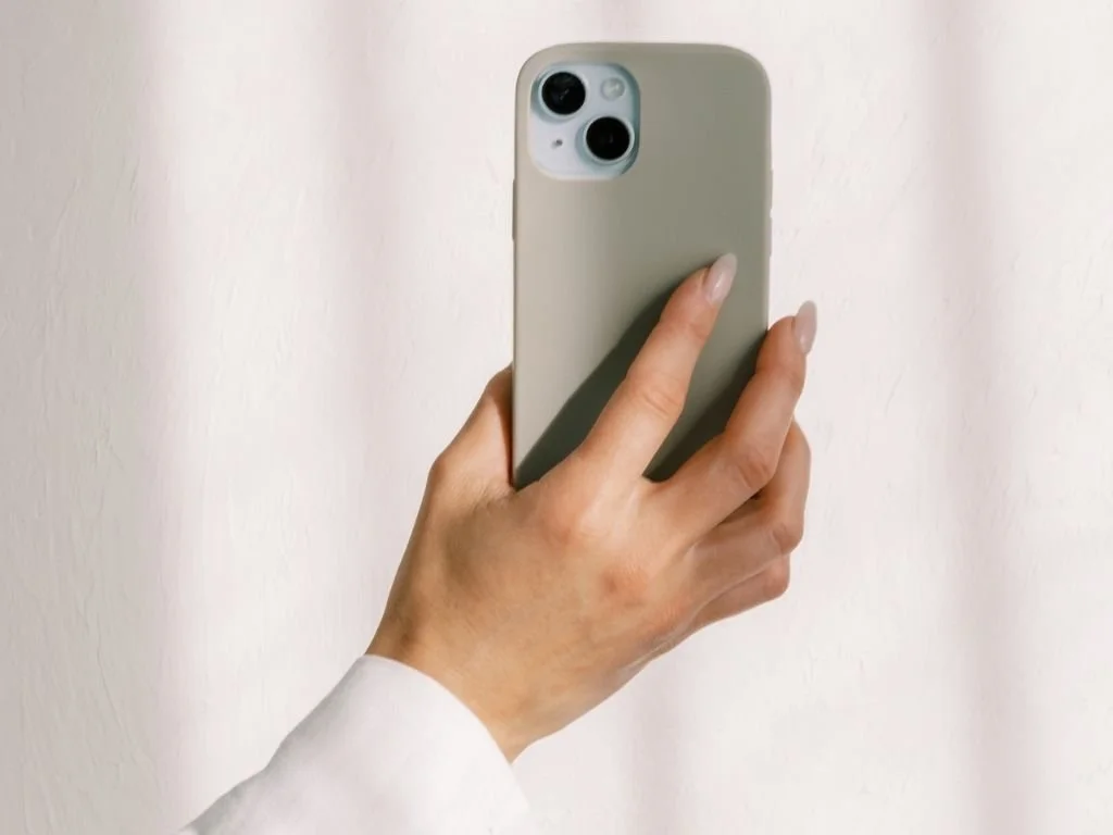 A person holding a smartphone with a beige case, taking a photo against a plain white wall.