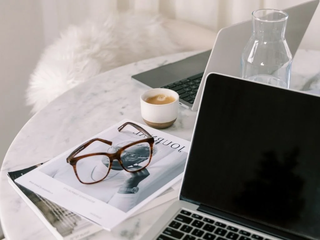 A marble table with a laptop, a cup of coffee, a glass water pitcher, a magazine, and a pair of glasses.