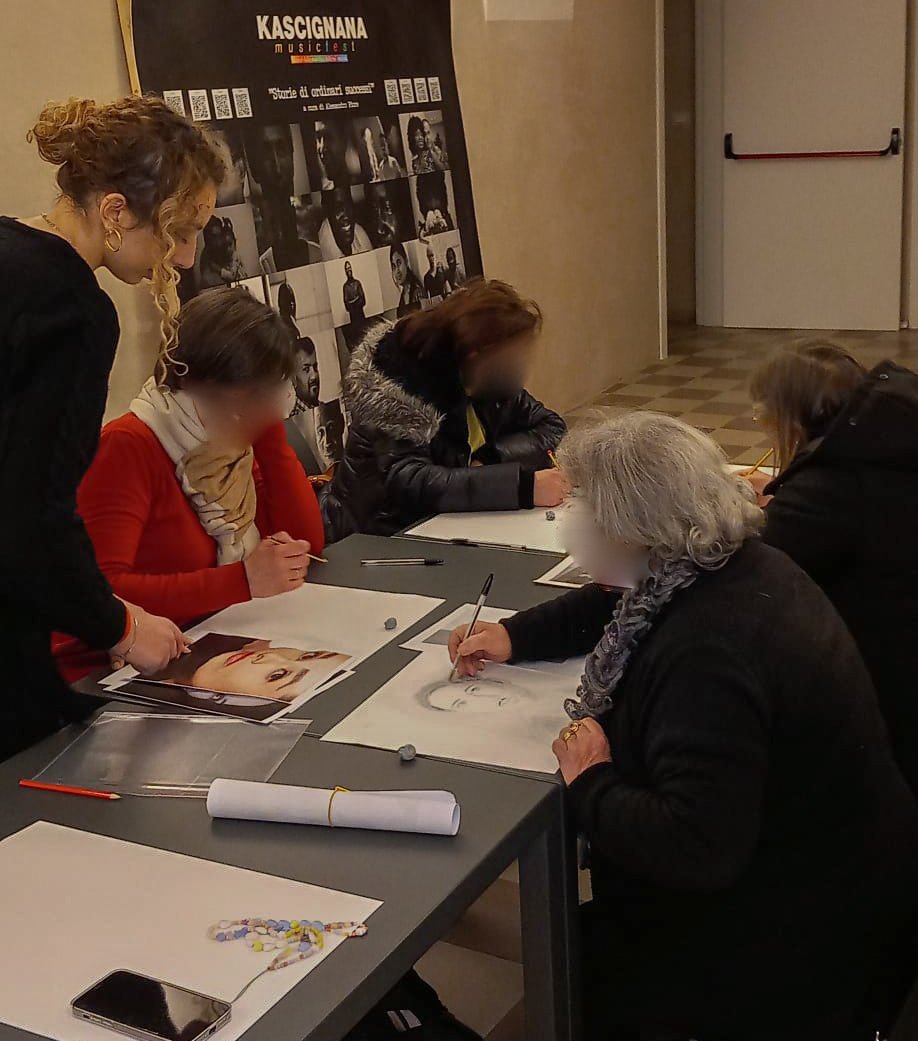 Gruppo di persone che disegnano e scrivono su fogli, intorno a un tavolo, con poster e fotografie sullo sfondo.