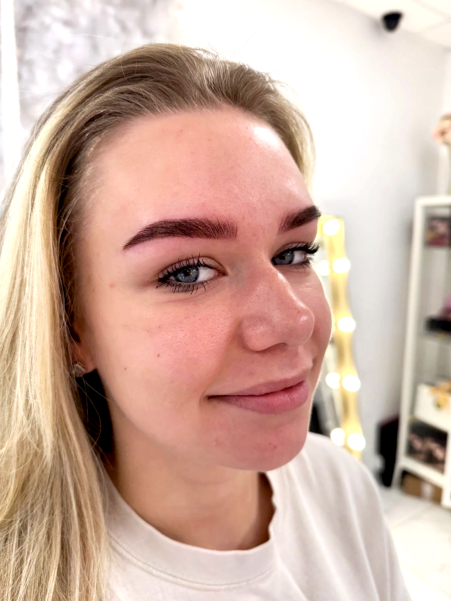 Close-up of a woman with blonde hair and blue eyes, showing her freshly done eyebrow makeup, in a room with a vanity mirror and mirror lights in the background.