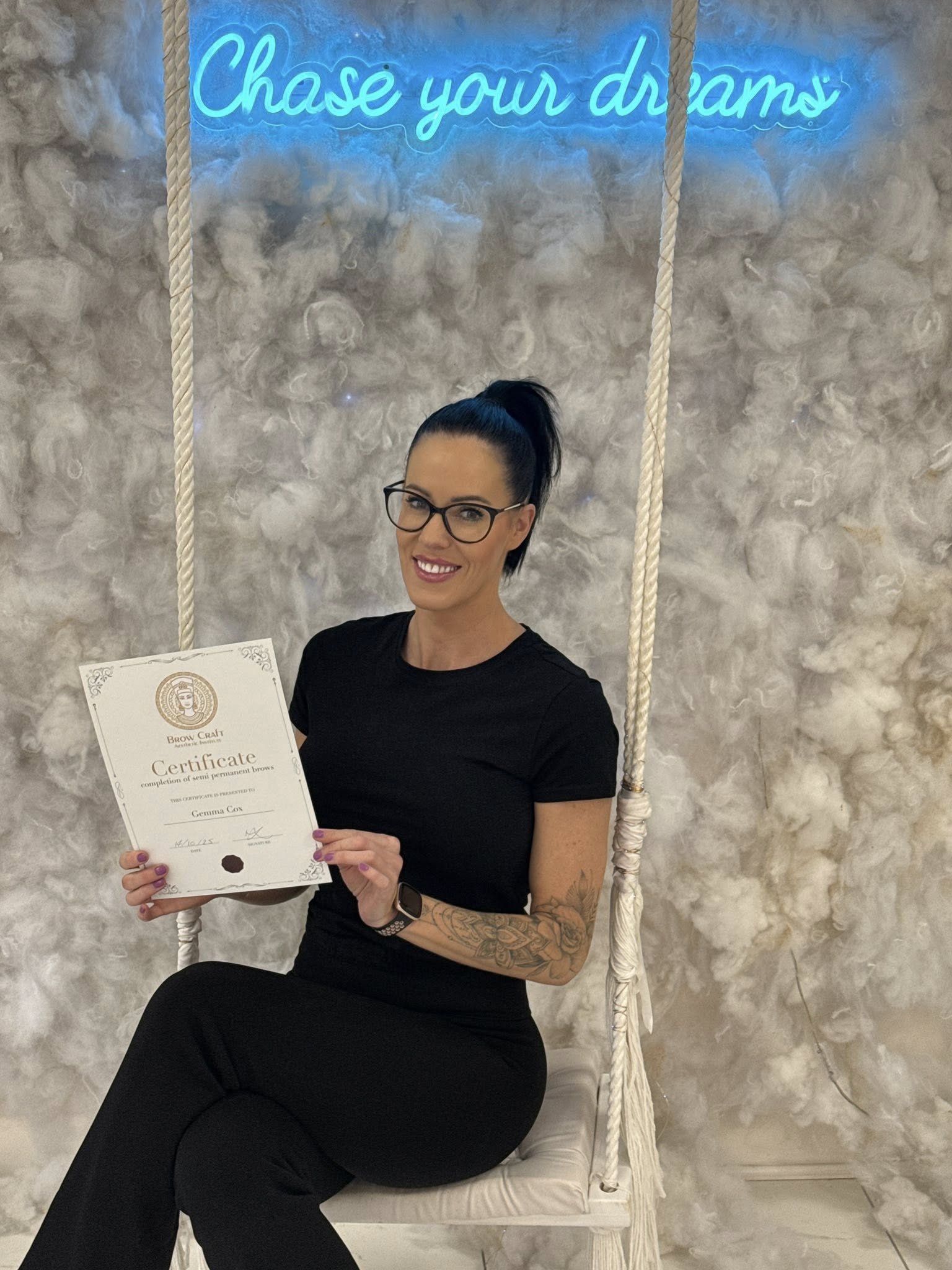 Woman with glasses sitting on a white swing holding a certificate. Behind her is a fluffy, cloud-like background with a blue neon sign that reads 'Chase your dreams'.