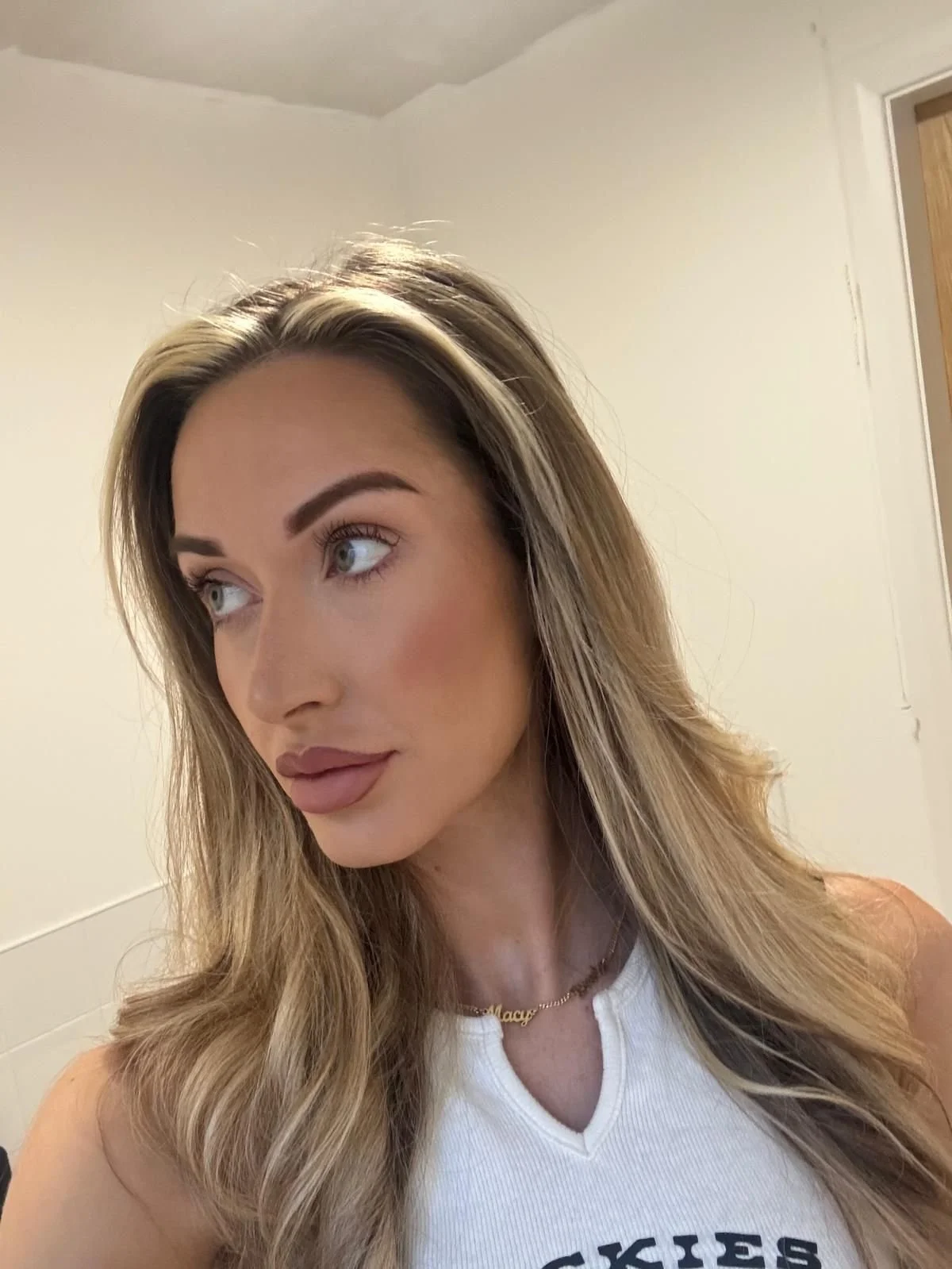 A woman with long, wavy blonde hair wearing a white sleeveless top and gold necklace, looking to her left in a room with beige walls.