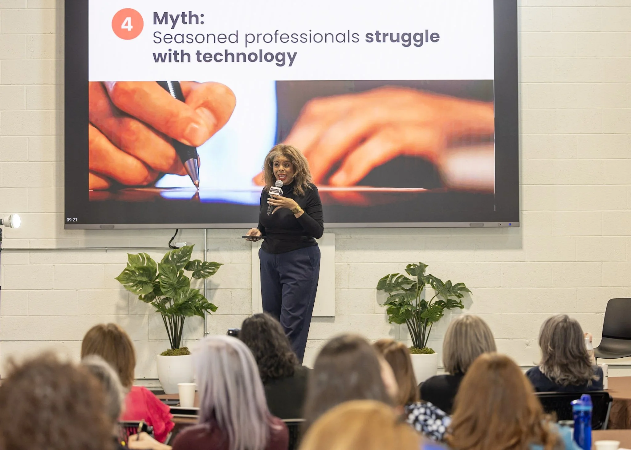 A woman is speaking to an audience in front of a large screen displaying a presentation slide. The slide has the title 'Myth: Seasoned professionals struggle with technology' and an image of a hand holding a pen. The audience seats are occupied by many people, and there are green potted plants in front of the speaker.