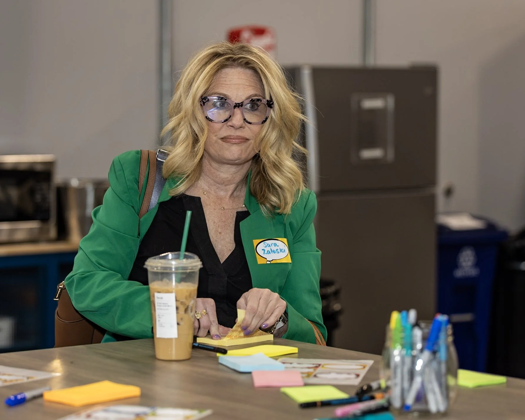A woman with blonde hair and glasses, wearing a green jacket, sitting at a table with a Starbucks drink, and various colorful sticky notes and pens in front of her, in what appears to be a meeting or workshop setting.
