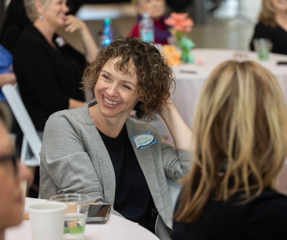 A woman with curly hair, wearing a gray blazer and a name tag that says 'Julie Snape,' is smiling and engaging in conversation at a social event or conference. Other women are also present, seated at tables with drinks and decorations.
