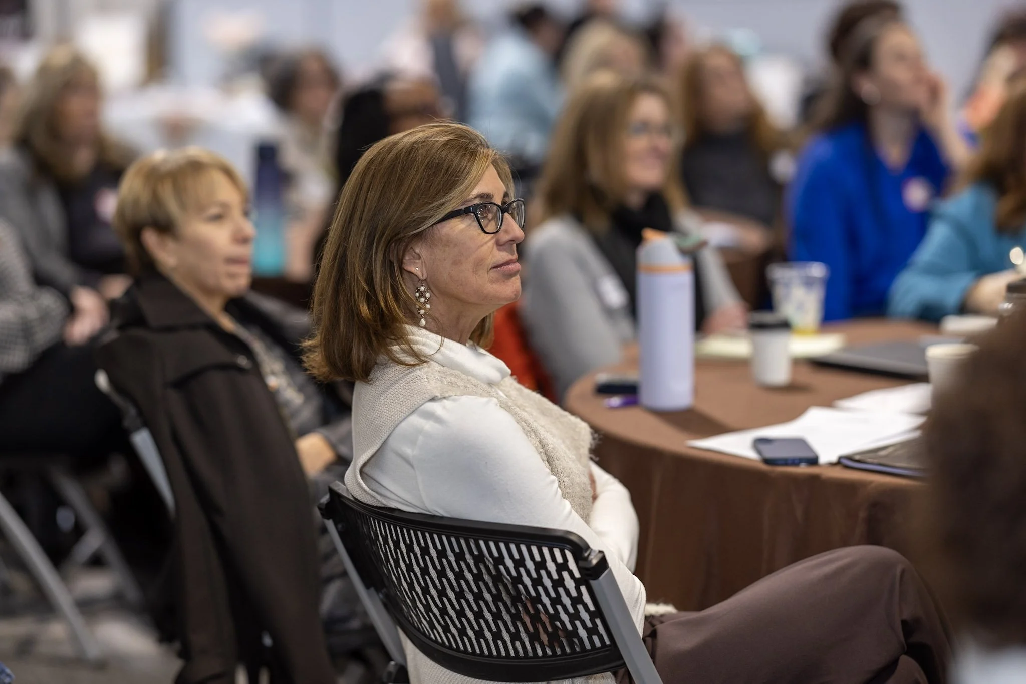 A woman with glasses and earrings sitting attentively at a conference or seminar, surrounded by other women also listening, with papers, a tablet, and a water bottle on the table.