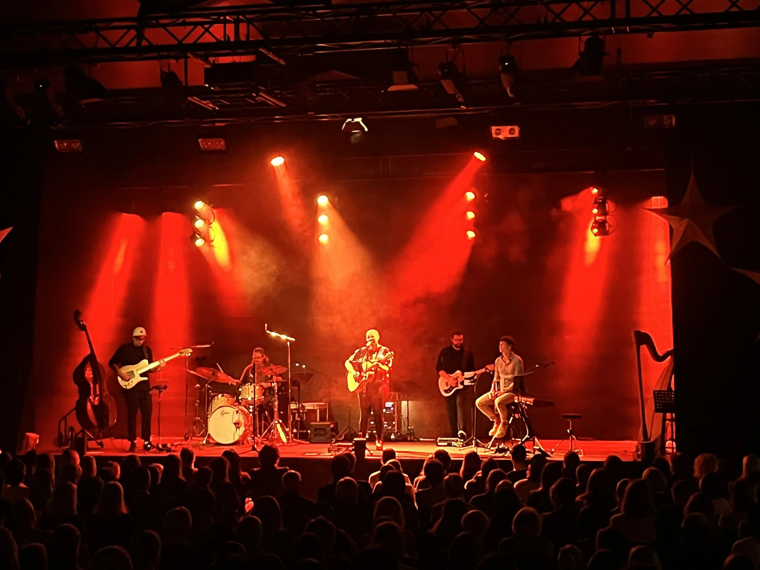 Eine Band spielt auf einer Bühne in einem dunklen Raum, beleuchtet von roten Scheinwerfern, während das Publikum im Vordergrund sitzt und zuschaut.