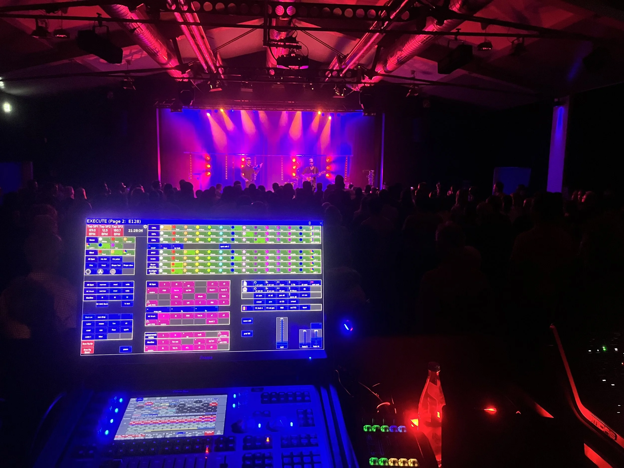 Eine dunkle Bühne mit bunten Lichtern, auf der eine Band auftritt. Im Vordergrund ist ein Computerbildschirm mit farbigen Steuertasten sichtbar.