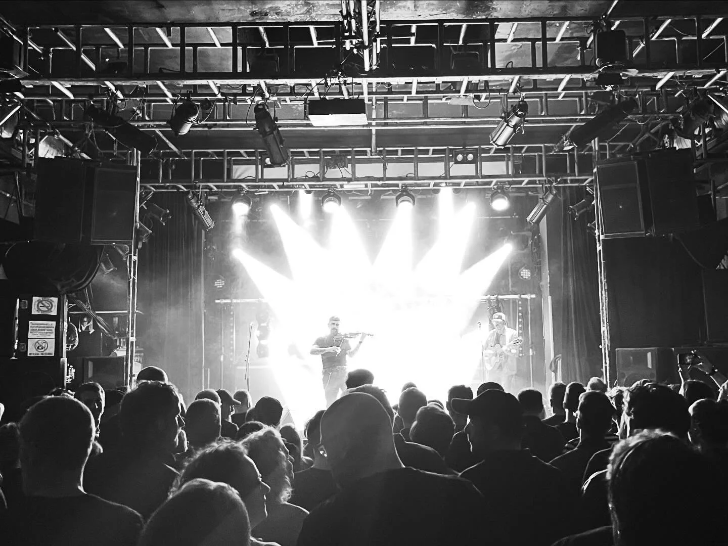 Konzerthalle mit Publikum im Vordergrund und Bühne im Hintergrund, auf der Musiker mit Instrumenten auftreten, bunte Lichter leuchten.