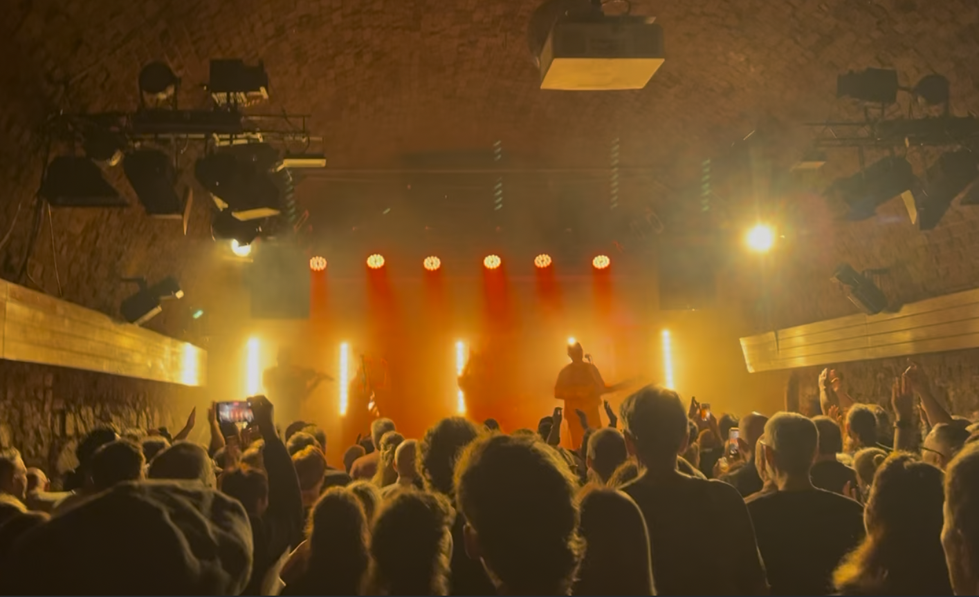 Eine Menschenmenge beobachtet eine Live-Musikperformance in einer dunklen, steinernen Location mit rotem und gelbem Bühnenlicht.