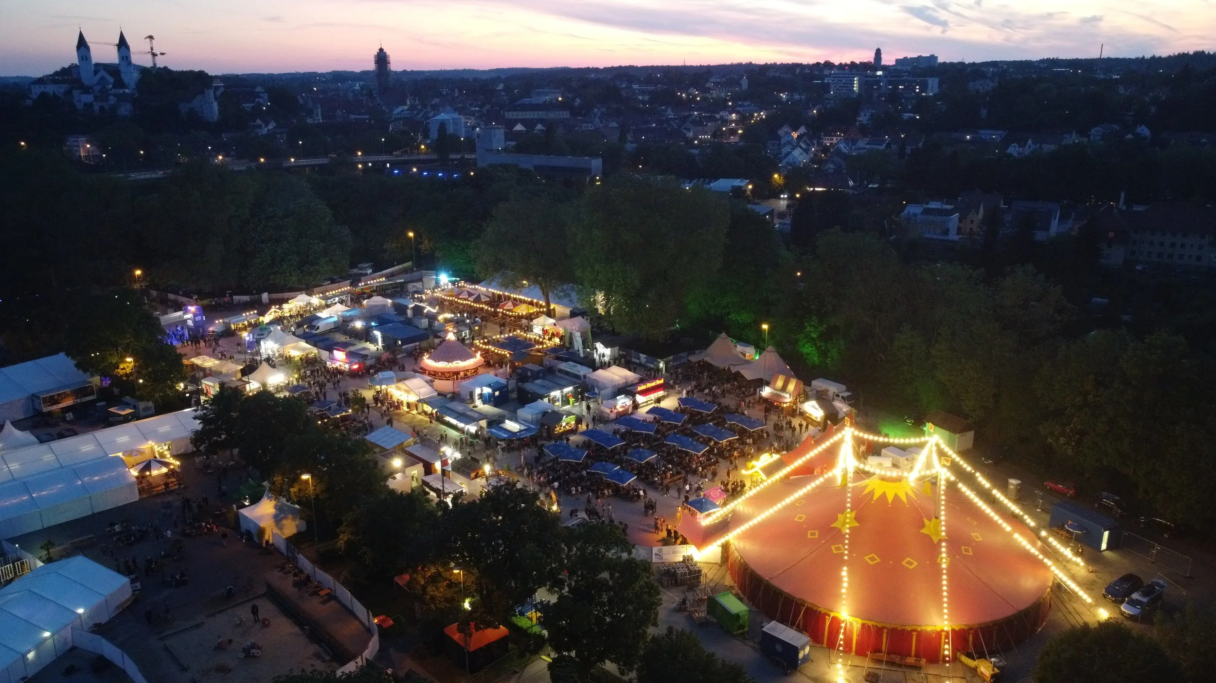 Abendliches Volksfest mit Lichterketten, Zelten, Karussell und Menschenmengen, Hintergrund Stadt mit Kirchen und Gebäuden, Uferlos Festival Freising