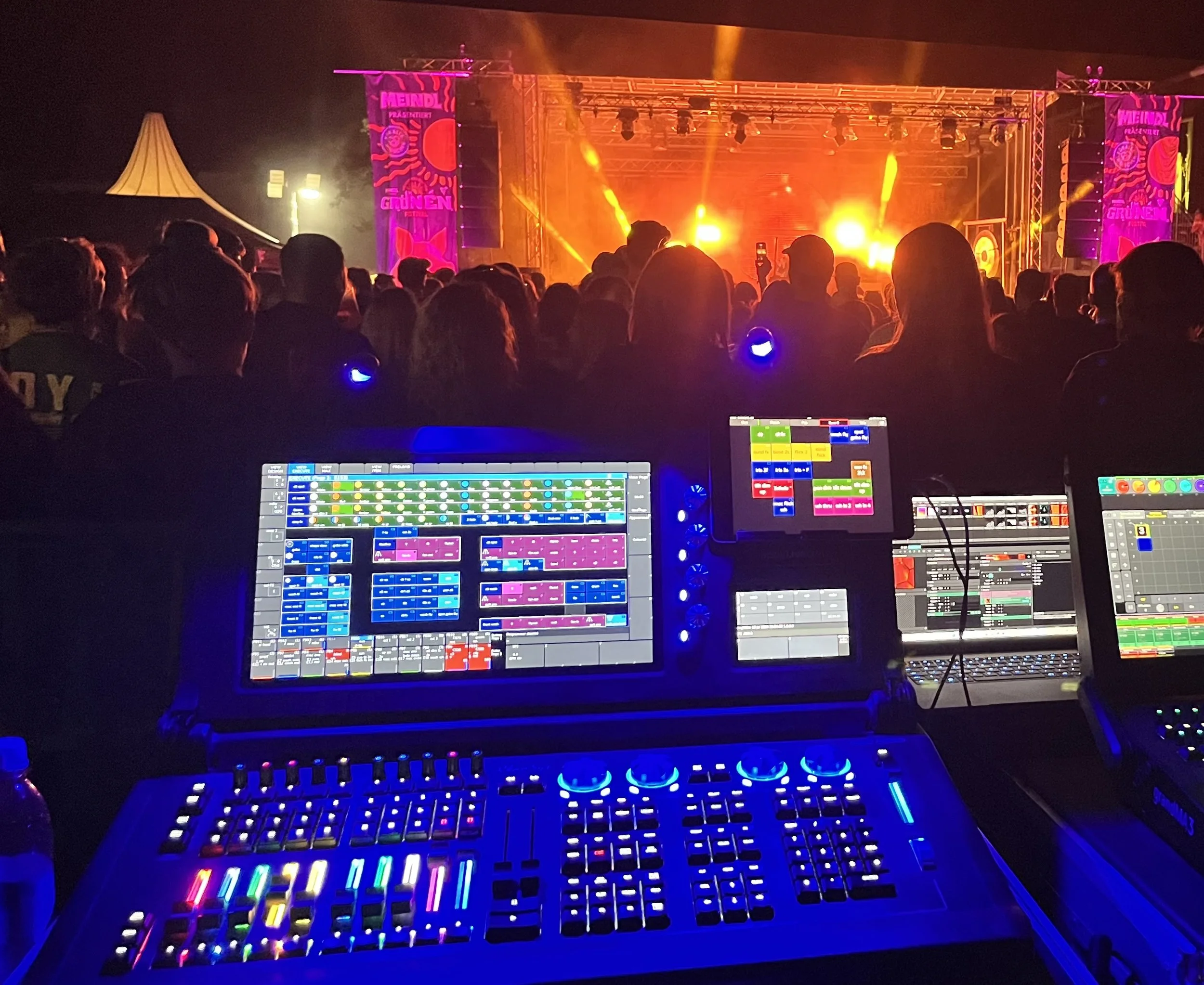 Auf einer Bühne sind bunte Lichter und Musikboxen zu sehen, während eine Menschenmenge zuschaut. Es sieht aus wie ein Konzert oder eine Veranstaltung bei Nacht.