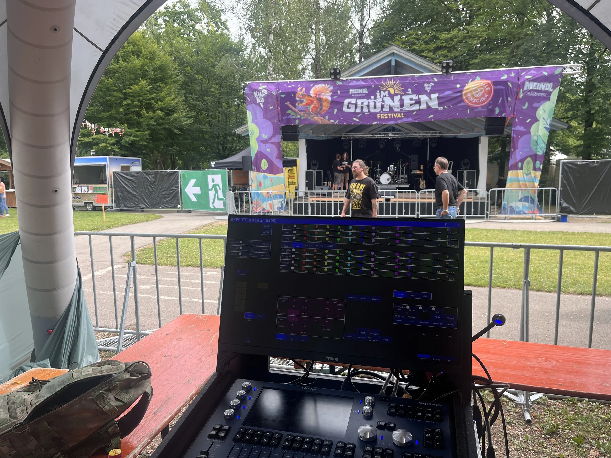 Bühne bei einem Festival im Freien mit einem Banner, auf dem 'Grünen Festival' steht, und einigen Menschen, die sich gerade unterhalten, im Hintergrund ist ein Baum.