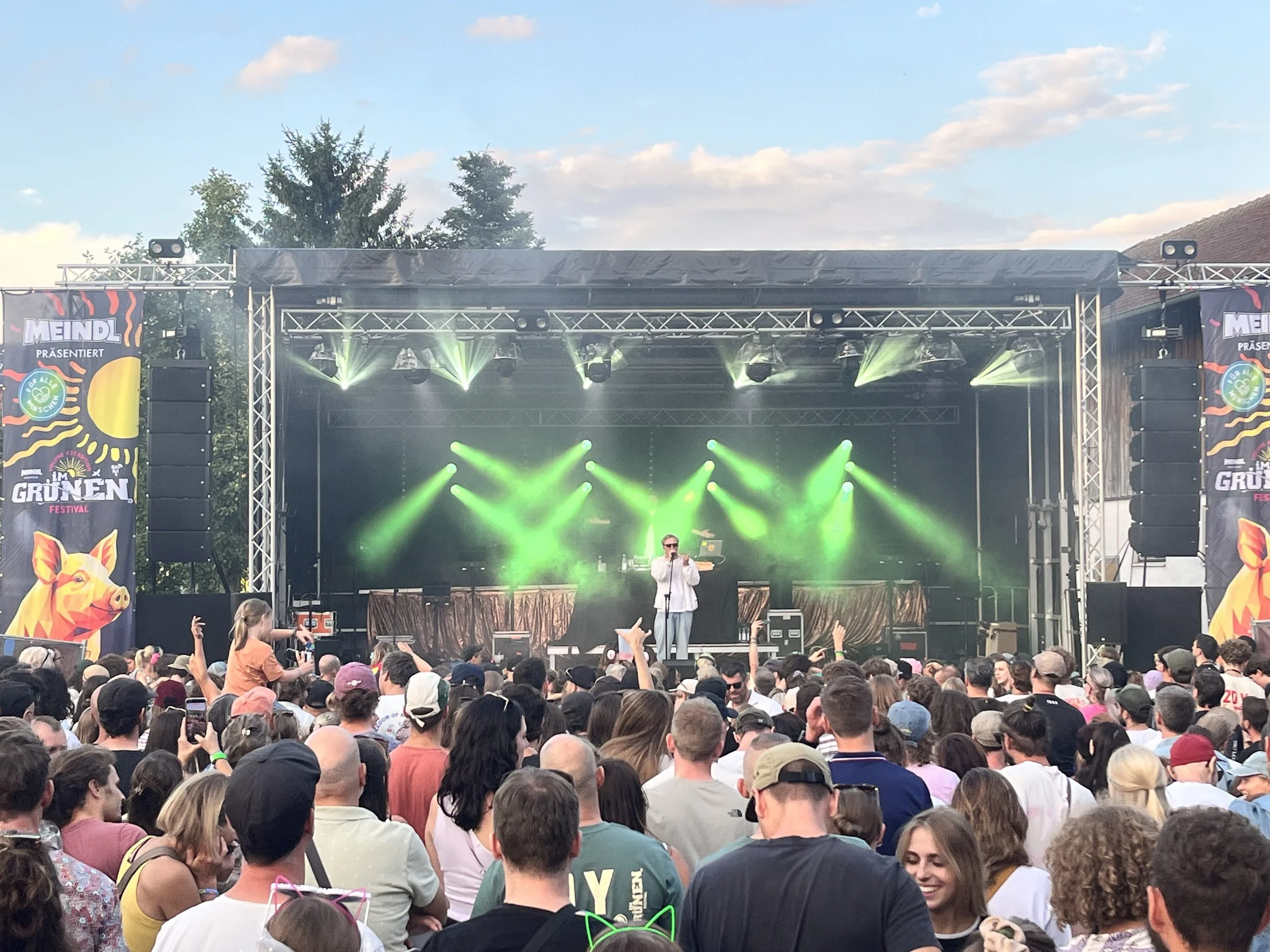 Ein großer Menschenauflauf vor einer Bühne bei einem Festival, mit Musikern, Lichtern und Banner mit Text auf der Bühne. im Grünen Festival 2026