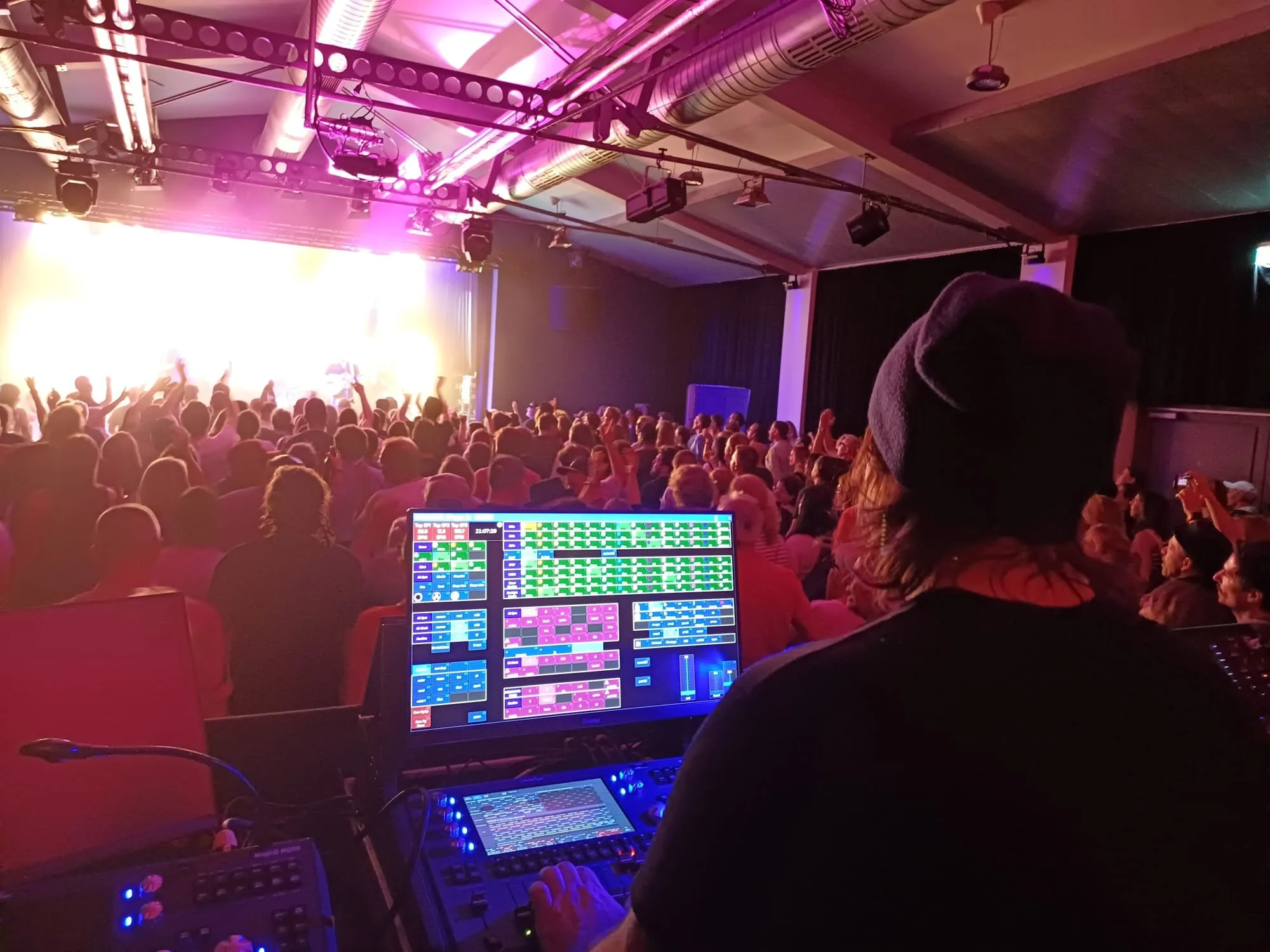 Der Blick auf die Bühne während eines Konzerts mit vielen Menschen im Publikum, eine Person an einem Tower mit Licht- und Tontechnik in einem Veranstaltungsraum.