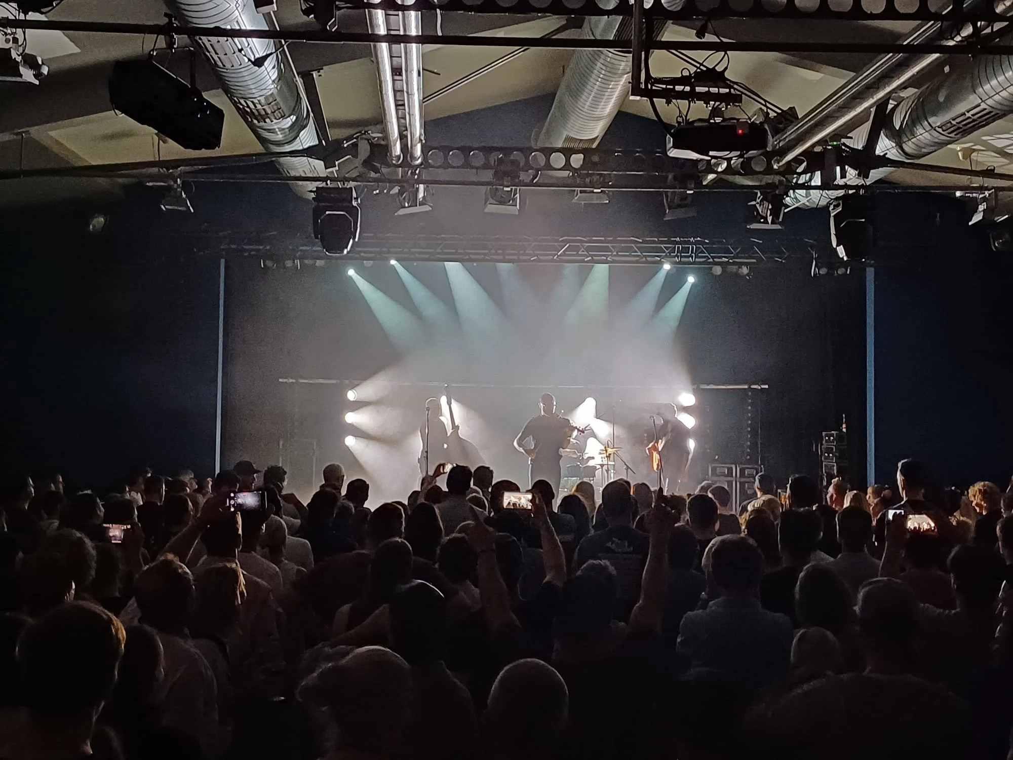 Blick auf eine Konzertbühne mit mehreren Musikern, Bühnenbeleuchtung und einer großen Menschenmenge im Publikum.Django 3000 mit happylivecrew Scene Wien 2026