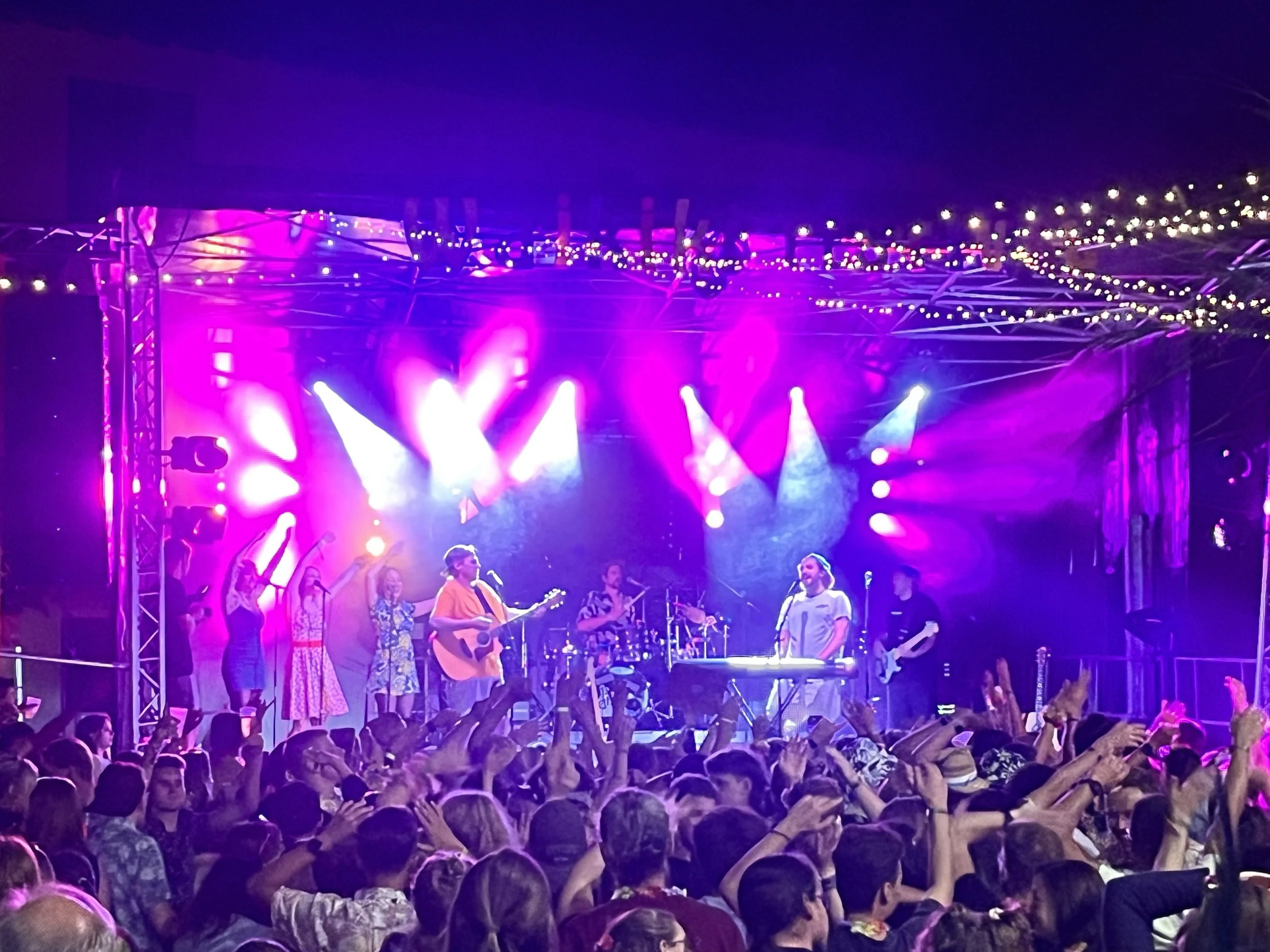 Menschen auf einer Bühne bei einem Konzert, mit Musiker, Gitarren und Keyboard, umgeben von bunten Lichteffekten, in einer großen Menschenmenge.
