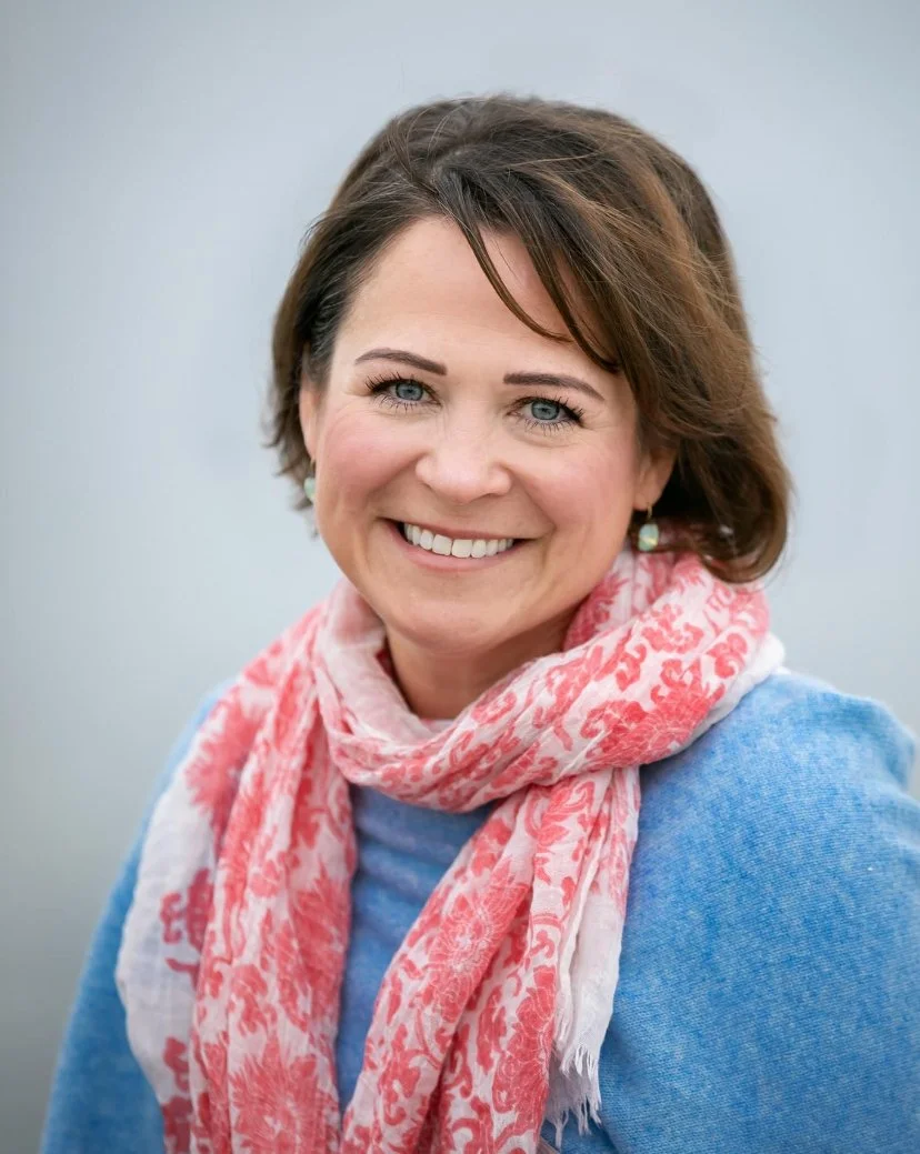 A woman with short brown hair smiling, wearing a light blue sweater and a pink and white patterned scarf, standing outdoors against a blurred gray background.