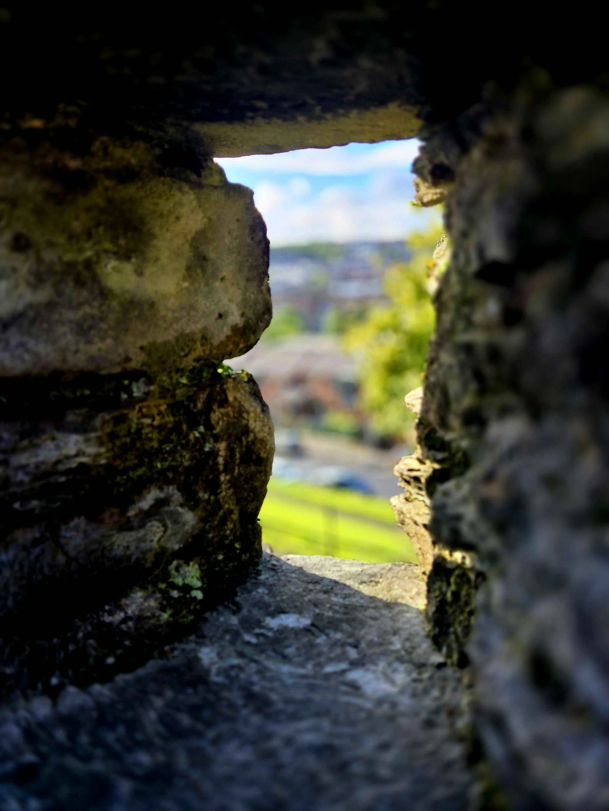 View through a narrow gap in a stone wall, showing a landscape with blue sky, white clouds, and green trees in the distance.