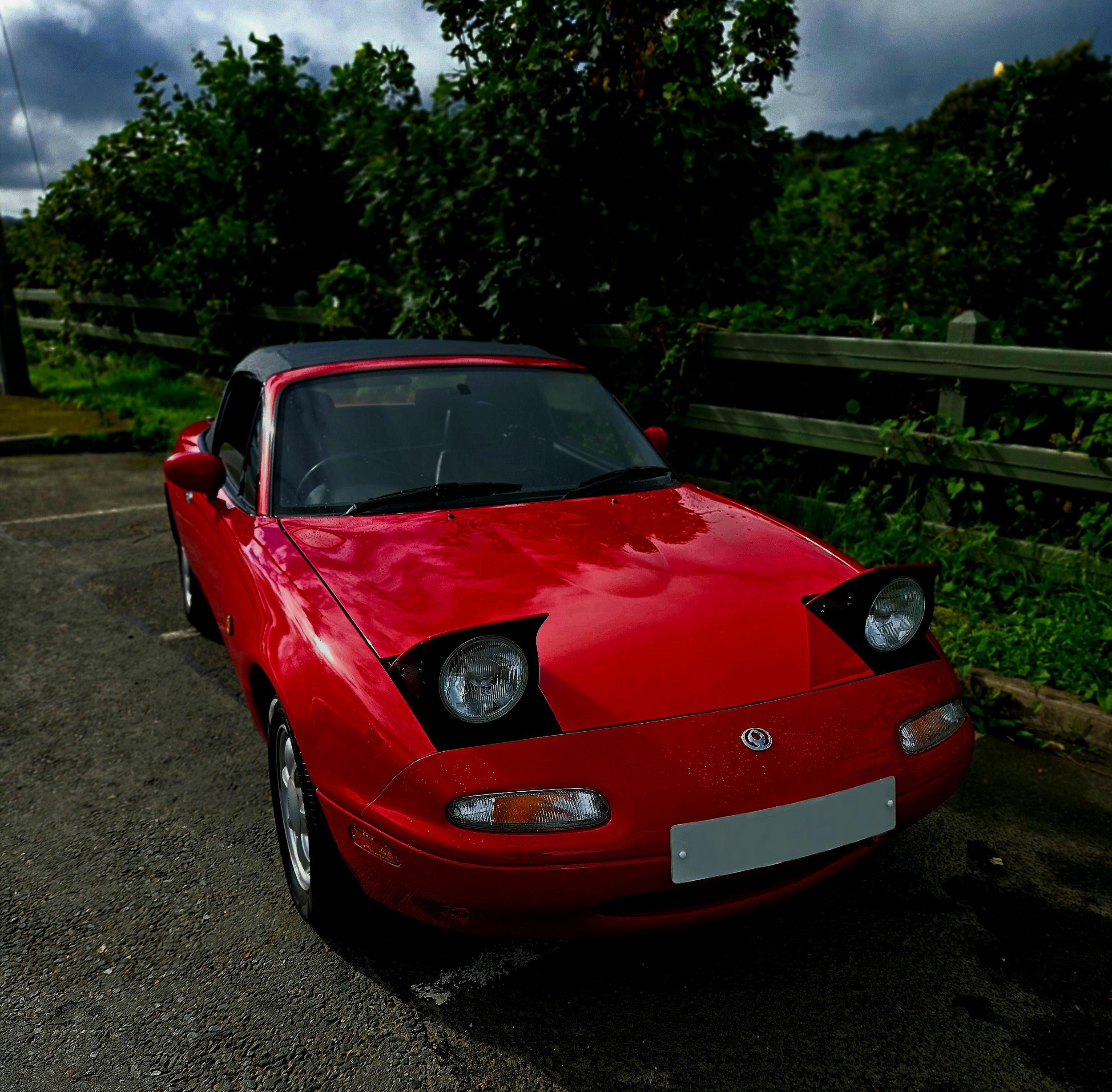 Red convertible sports car parked on a gravel lot near green bushes and trees, with a cloudy sky overhead.