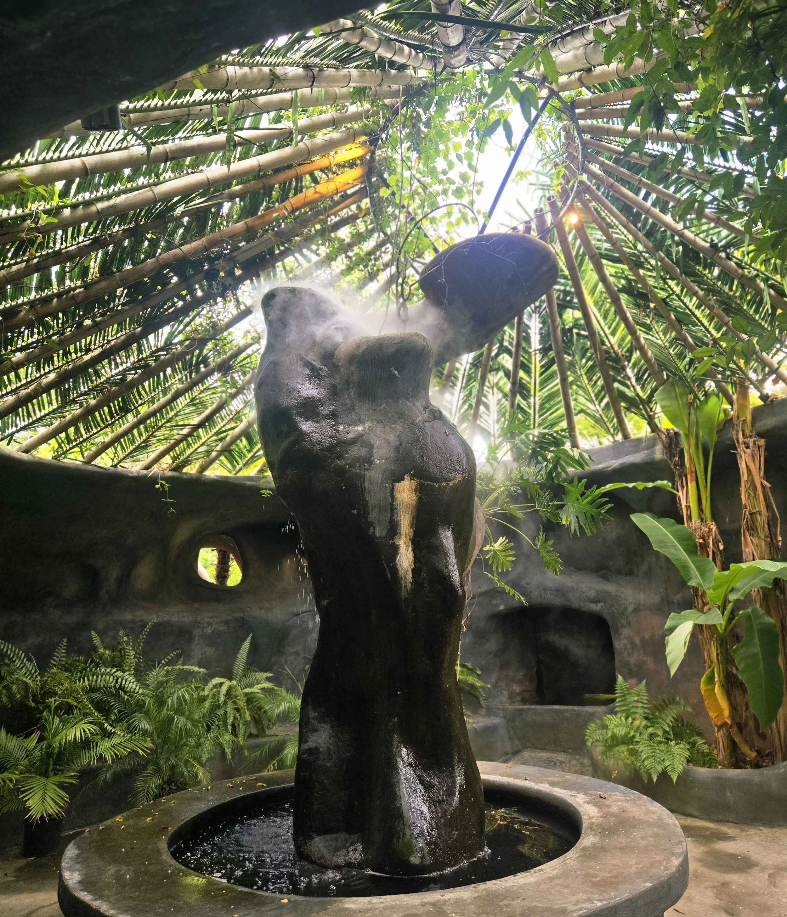 A steam rising black volcanic stone sculpture of a woman at the center of lush tropical vegetation, with bamboo and leafy plants surrounding the scene.