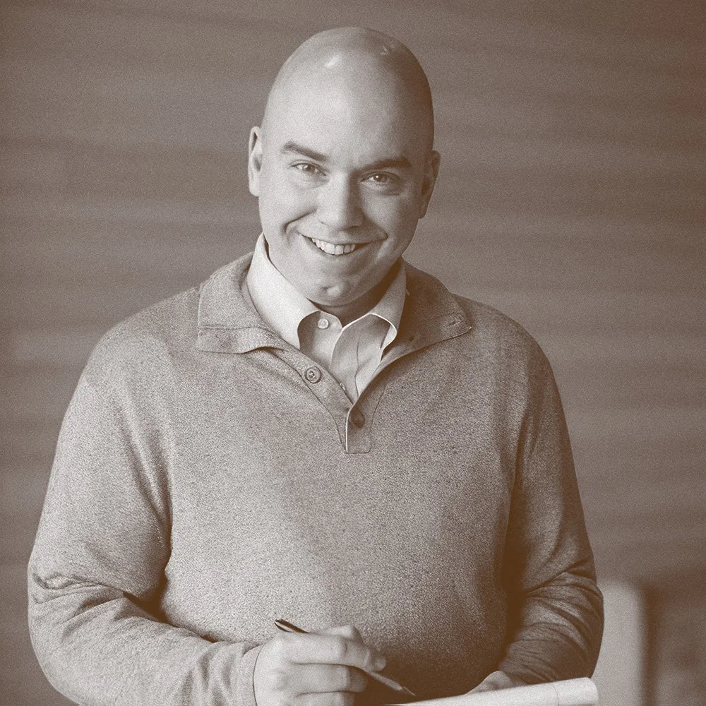 A smiling man holding a pen and clipboard, wearing a collared shirt and sweater, standing indoors.