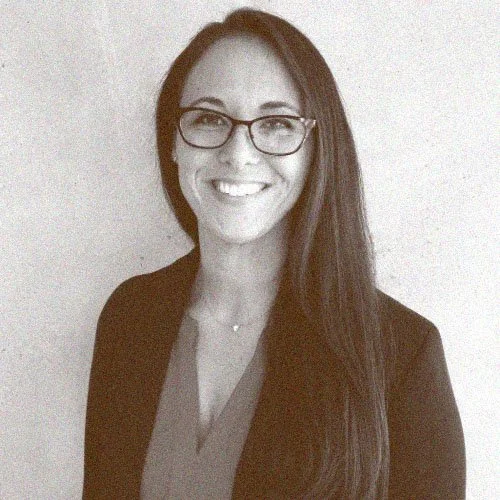 A woman with long dark hair and glasses smiling at the camera, dressed in a blazer and top against a plain background.
