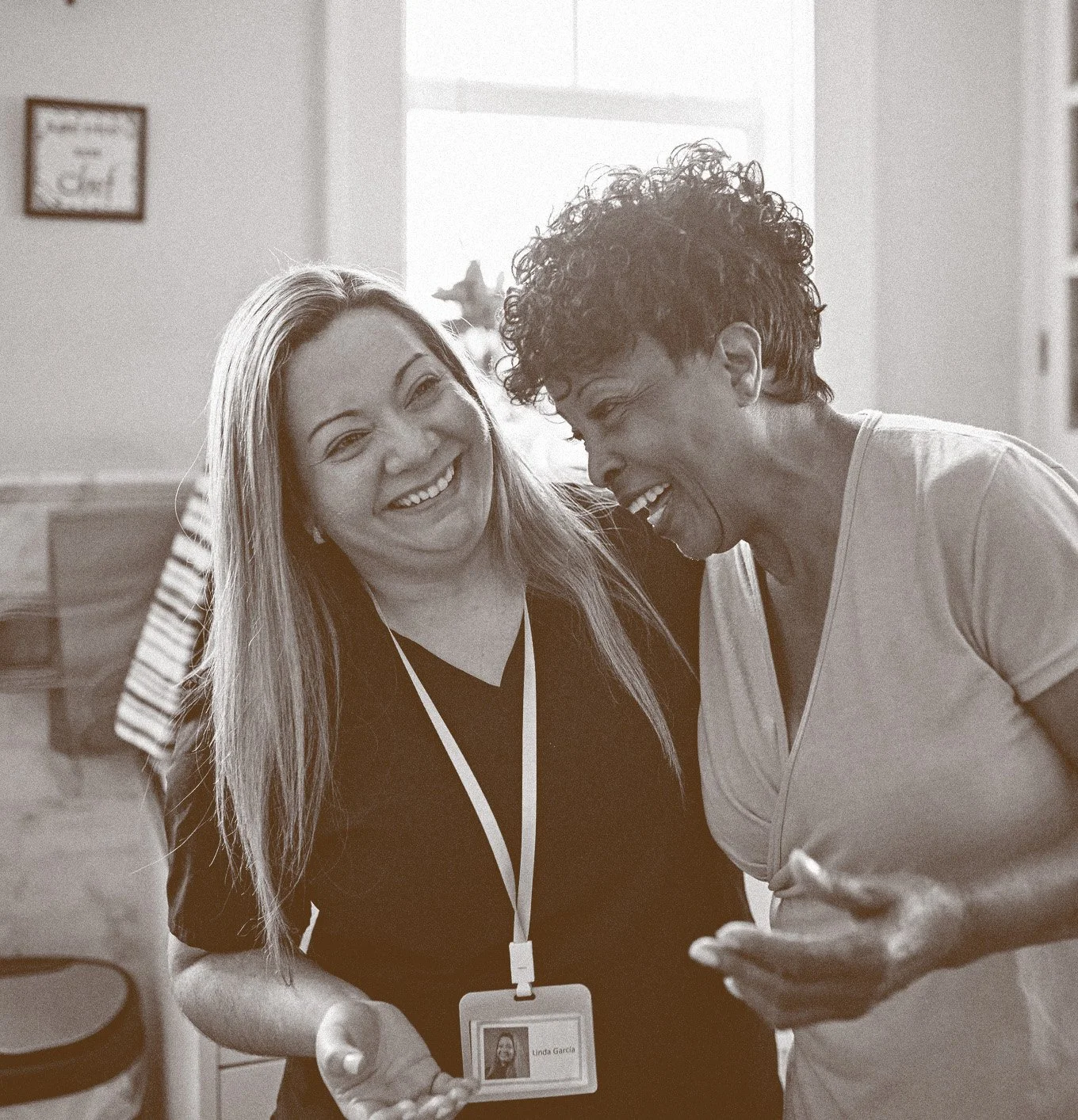 Two women smiling and laughing together indoors, one wearing a badge around her neck, the other holding a smartphone.
