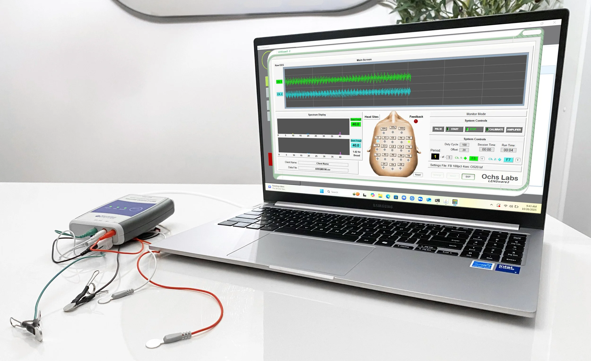A laptop displaying neurofeedback software connected to a small medical device with attached electrode leads on a white desk.