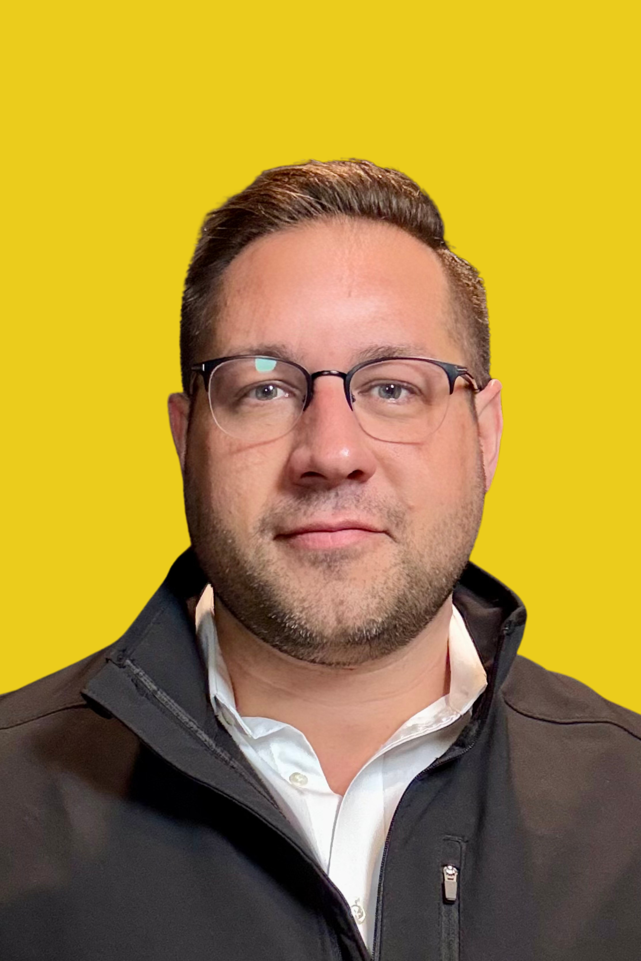 A man with glasses, short brown hair, and a beard wearing a white shirt and black jacket, posed against a yellow background.