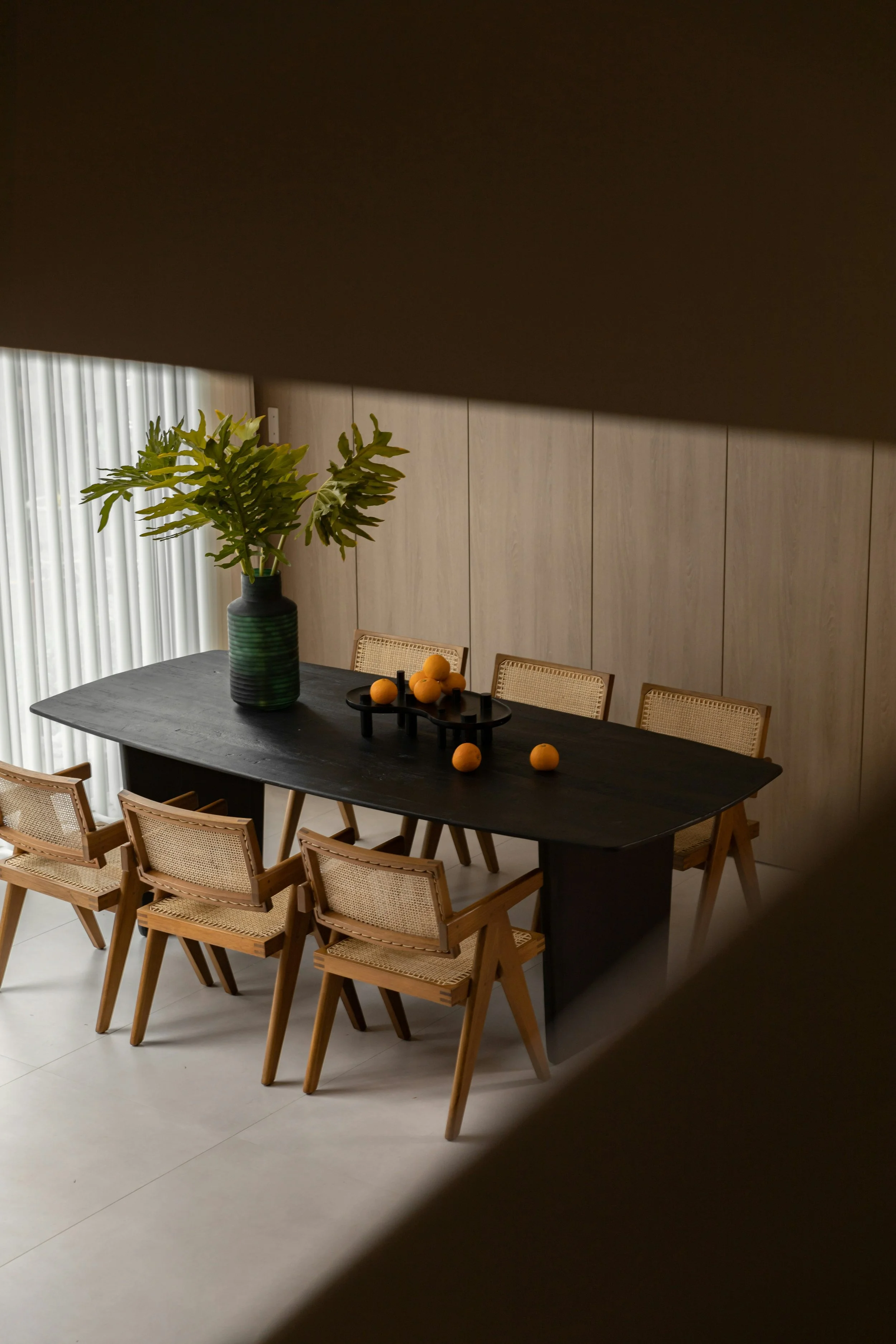 Dining room with a black table, six wooden chairs, a large green potted plant, and yellow citrus fruits on the table.