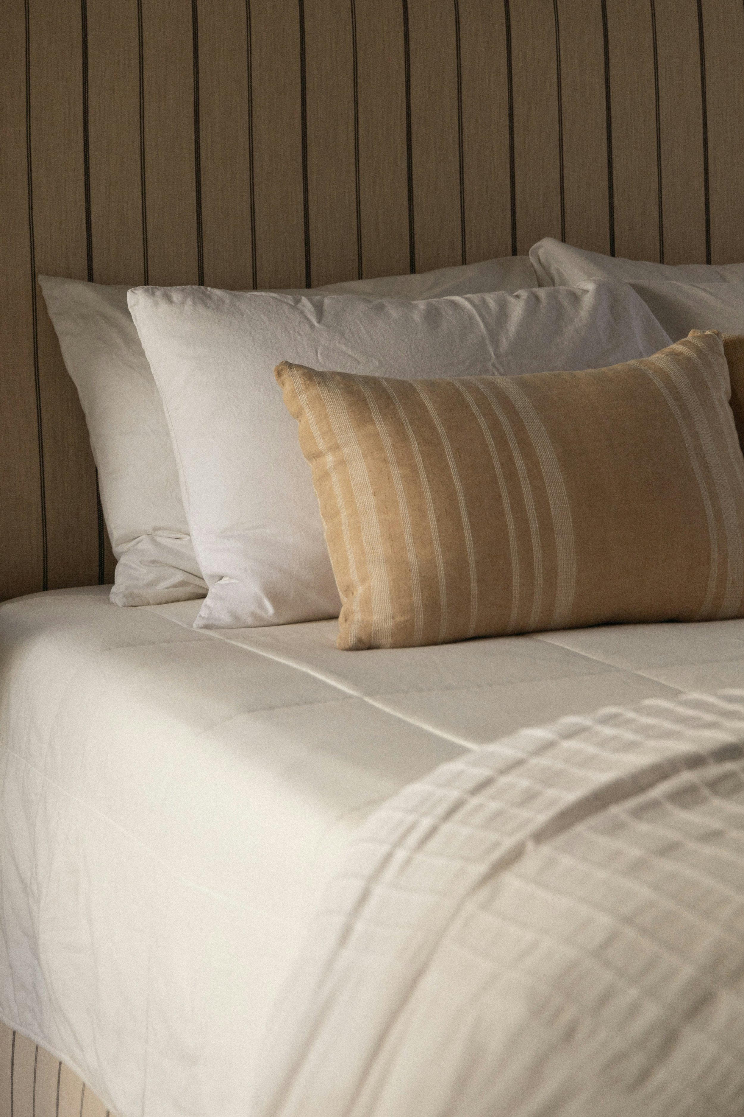 A neatly made bed with white pillows, a beige decorative pillow, and a cream-colored bedspread, against a wooden paneled wall.