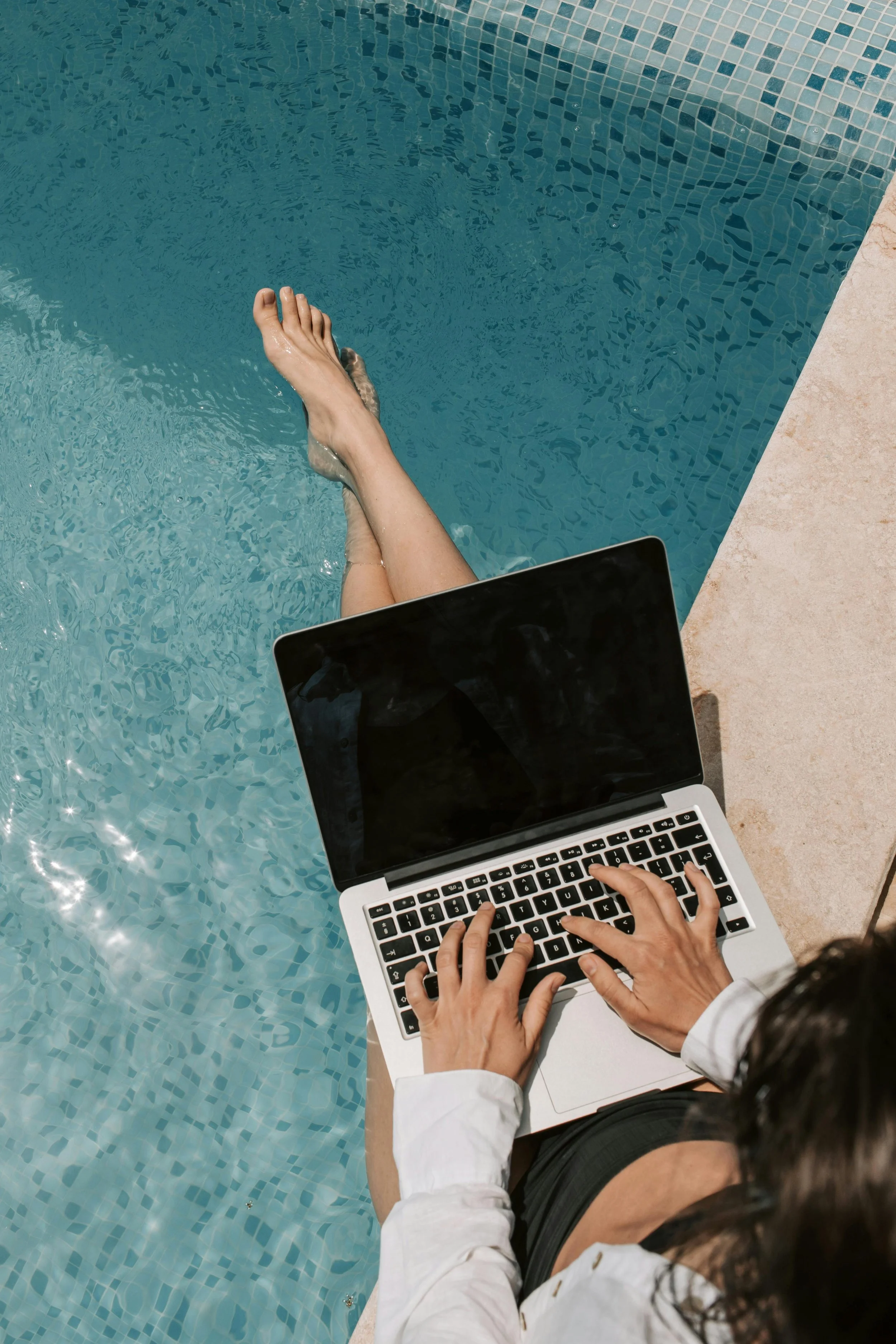 A woman sitting on a cushion by a pool, working on a laptop, with tropical plants and a glass wall in the background.