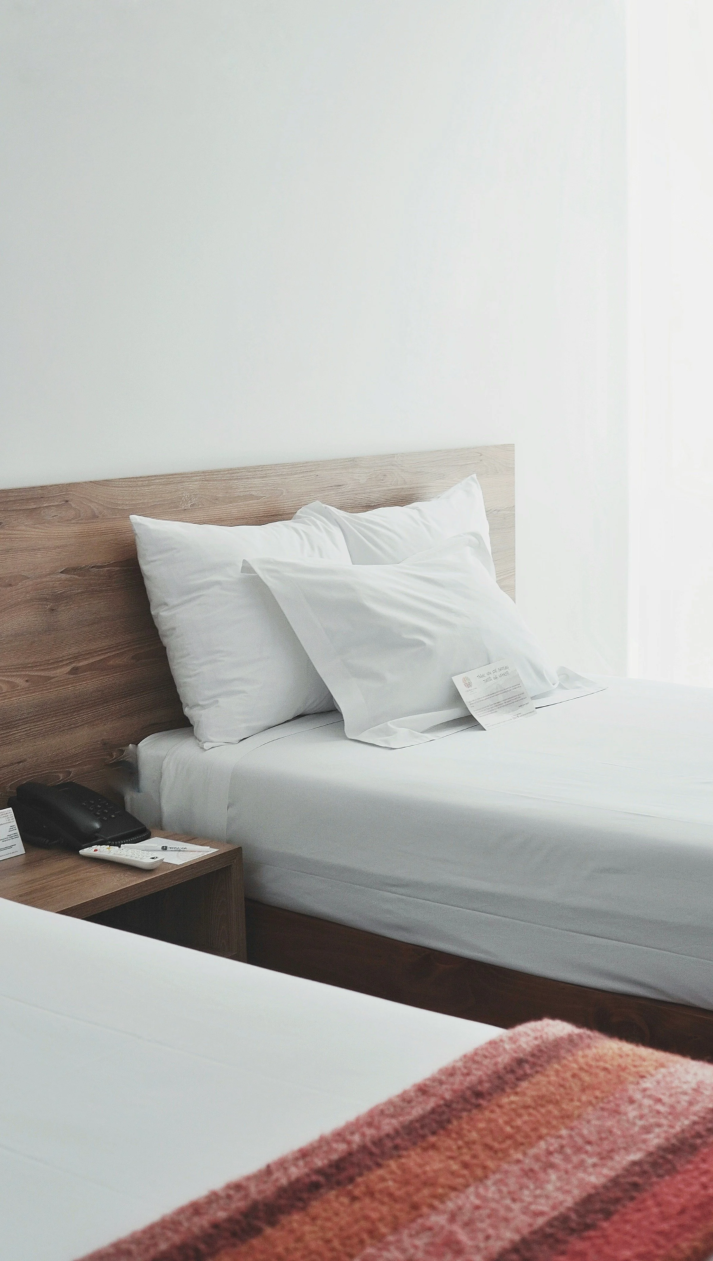 A hotel room with a made bed, white pillows, a wooden headboard, a nightstand with a phone, remote, and paper, and part of a second bed with a colorful blanket.