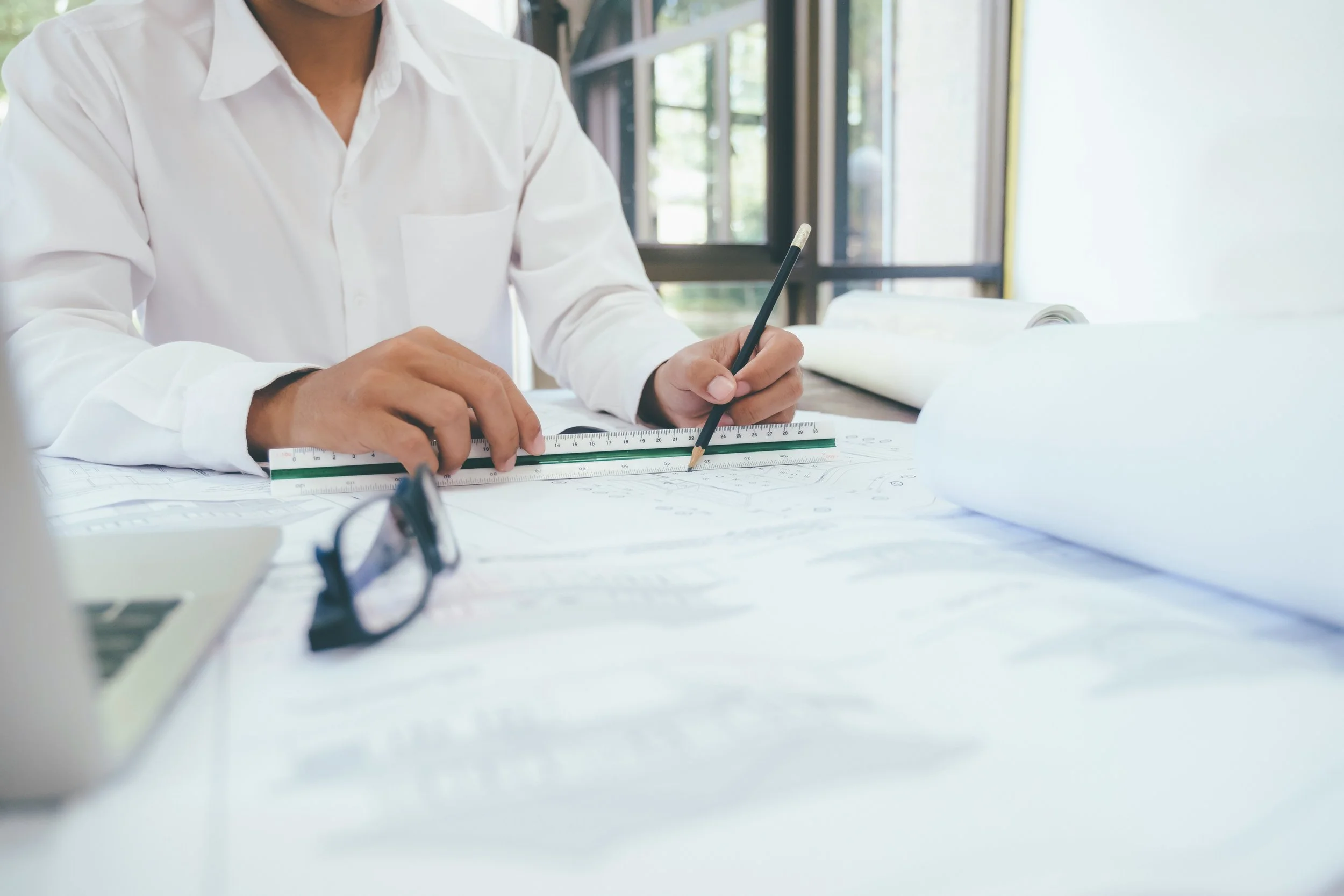An architect in a white shirt using a ruler and pencil to plan on blueprints, with glasses and a laptop nearby in a bright office.