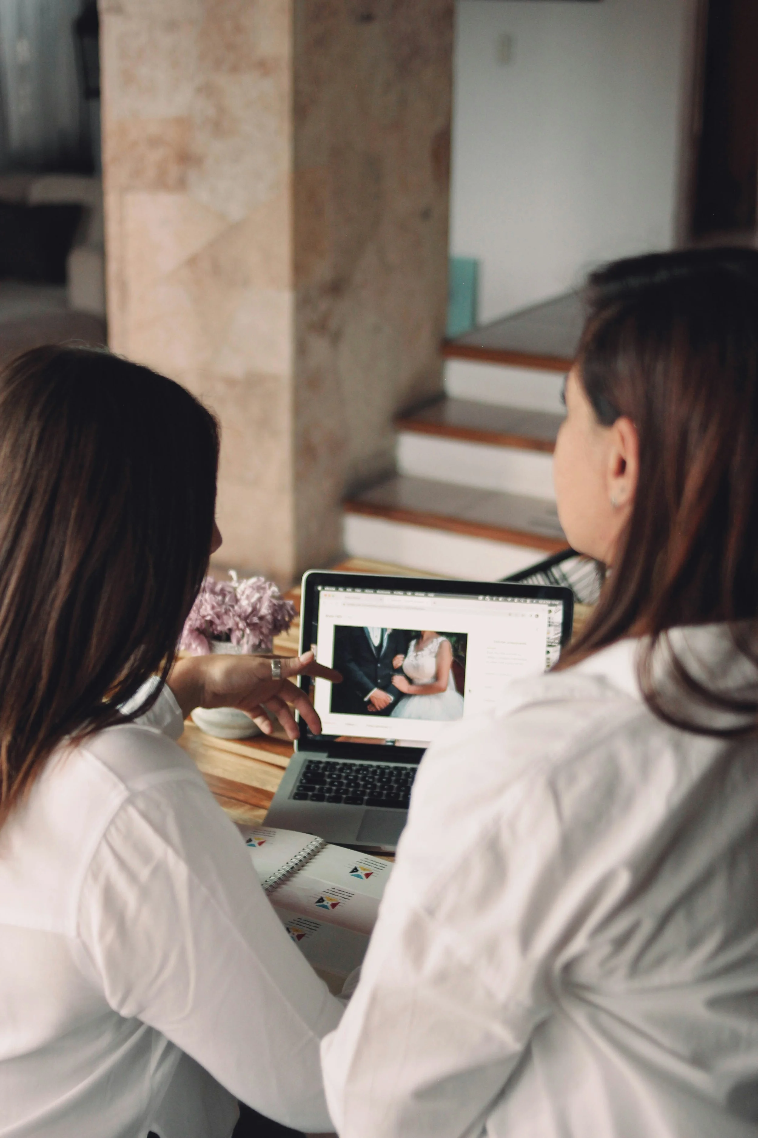 Two women sitting at a table, looking at a photo of a couple in wedding attire on a laptop computer.