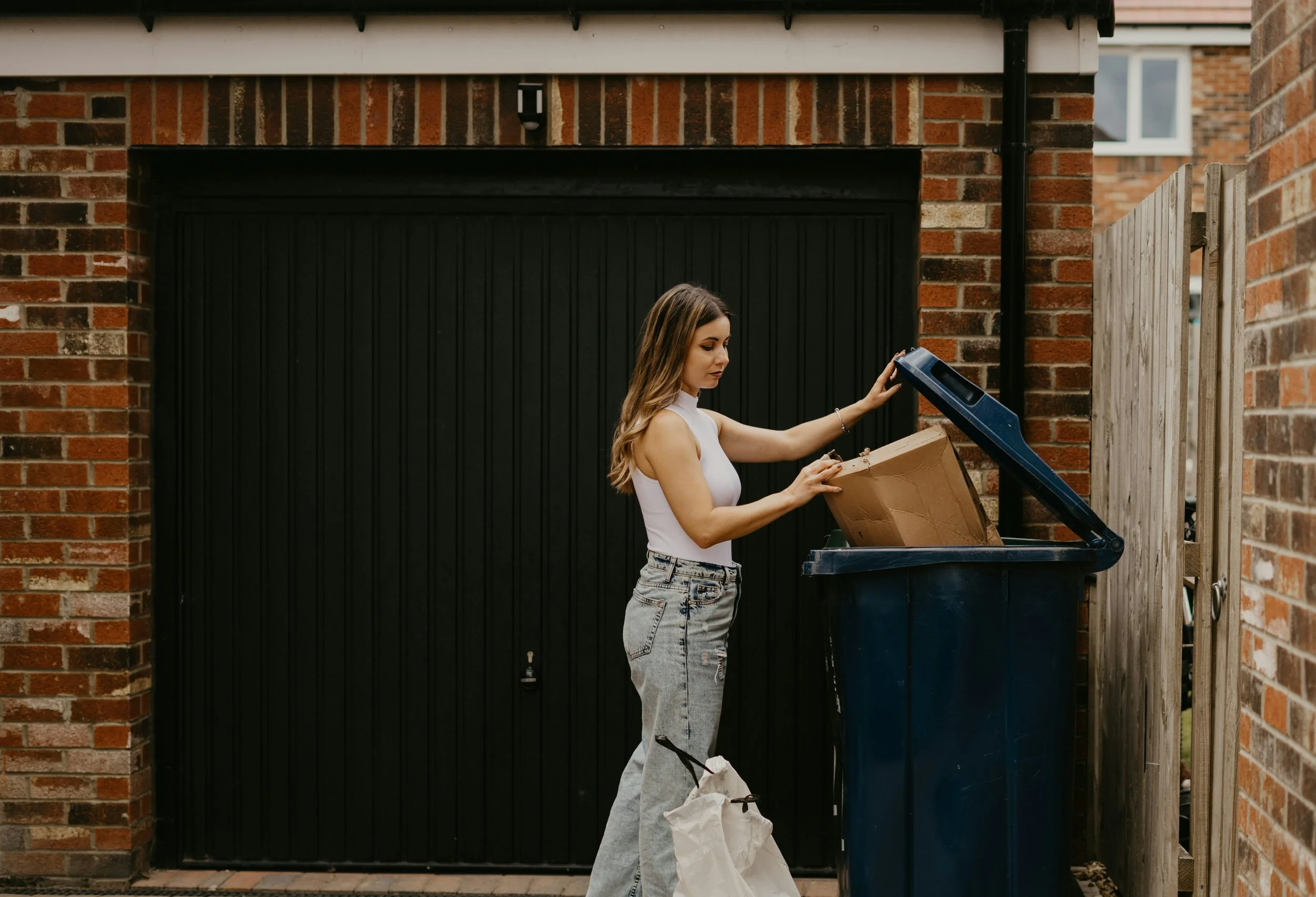 A young woman with long hair wearing a white tank top and ripped jeans is standing next to a blue trash bin with the lid open, looking inside, outside a brick building with a black garage door and a wooden fence.
