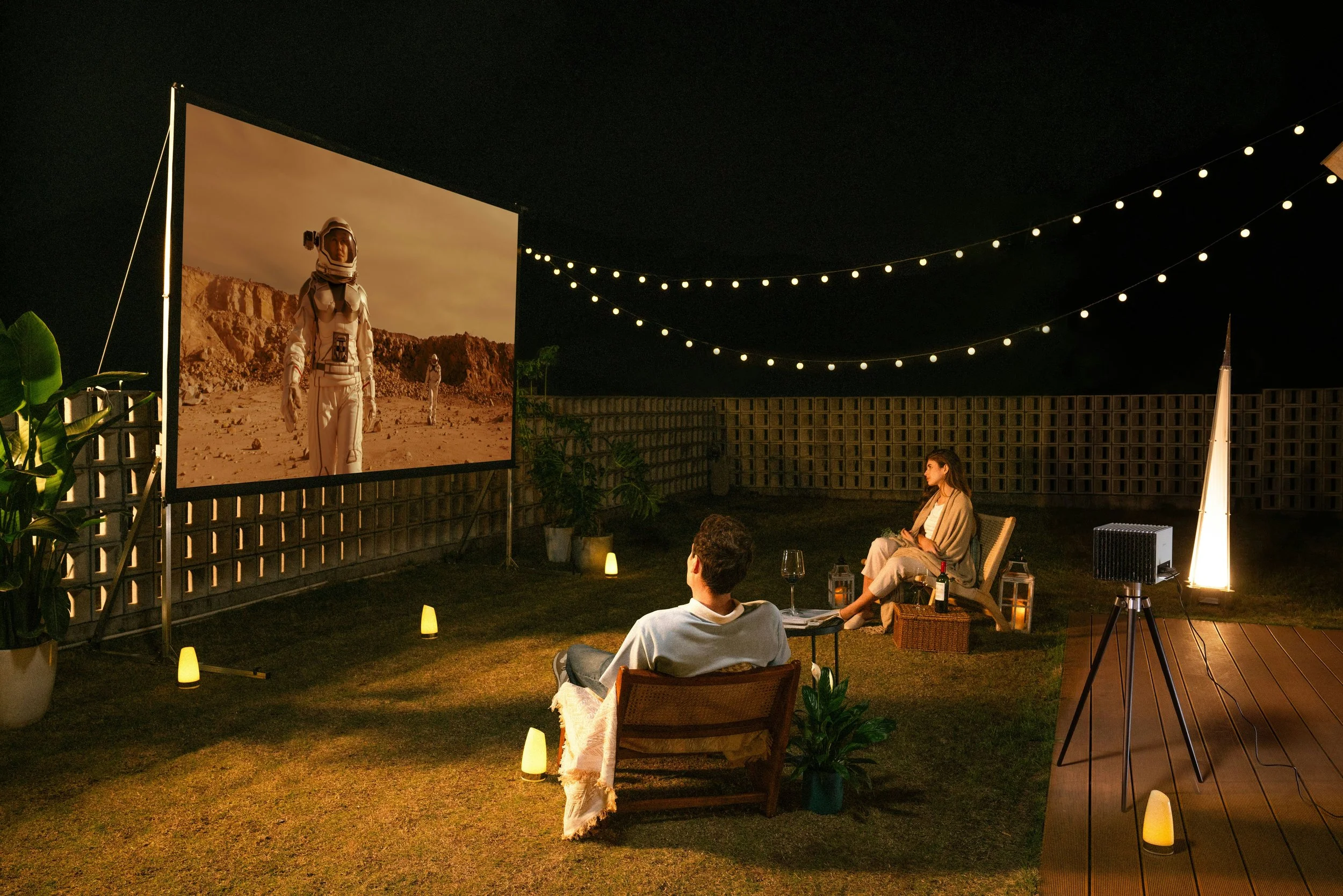 A couple watching a movie outdoors at night on a large screen with astronaut scenes, in a backyard with string lights, potted plants, lanterns, a coffee table with drinks and snacks, and a projector.